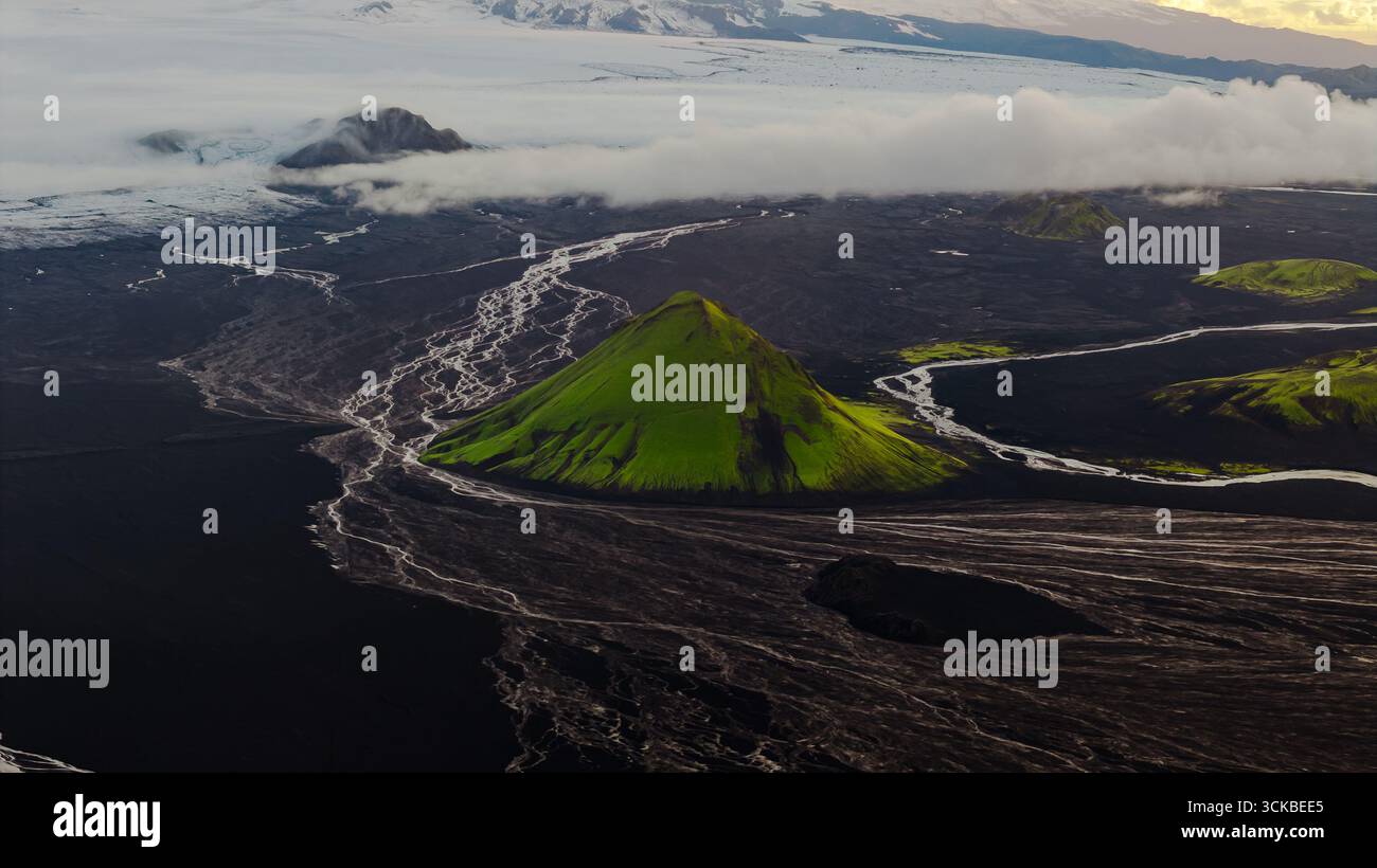 Aus der Vogelperspektive eines grünen Vulkankegels inmitten dunkler Ebenen mit weißen Flüssen, einem Gletscher und schneebedeckten Gipfeln unter niedrigen Wolken in Island. Stockfoto