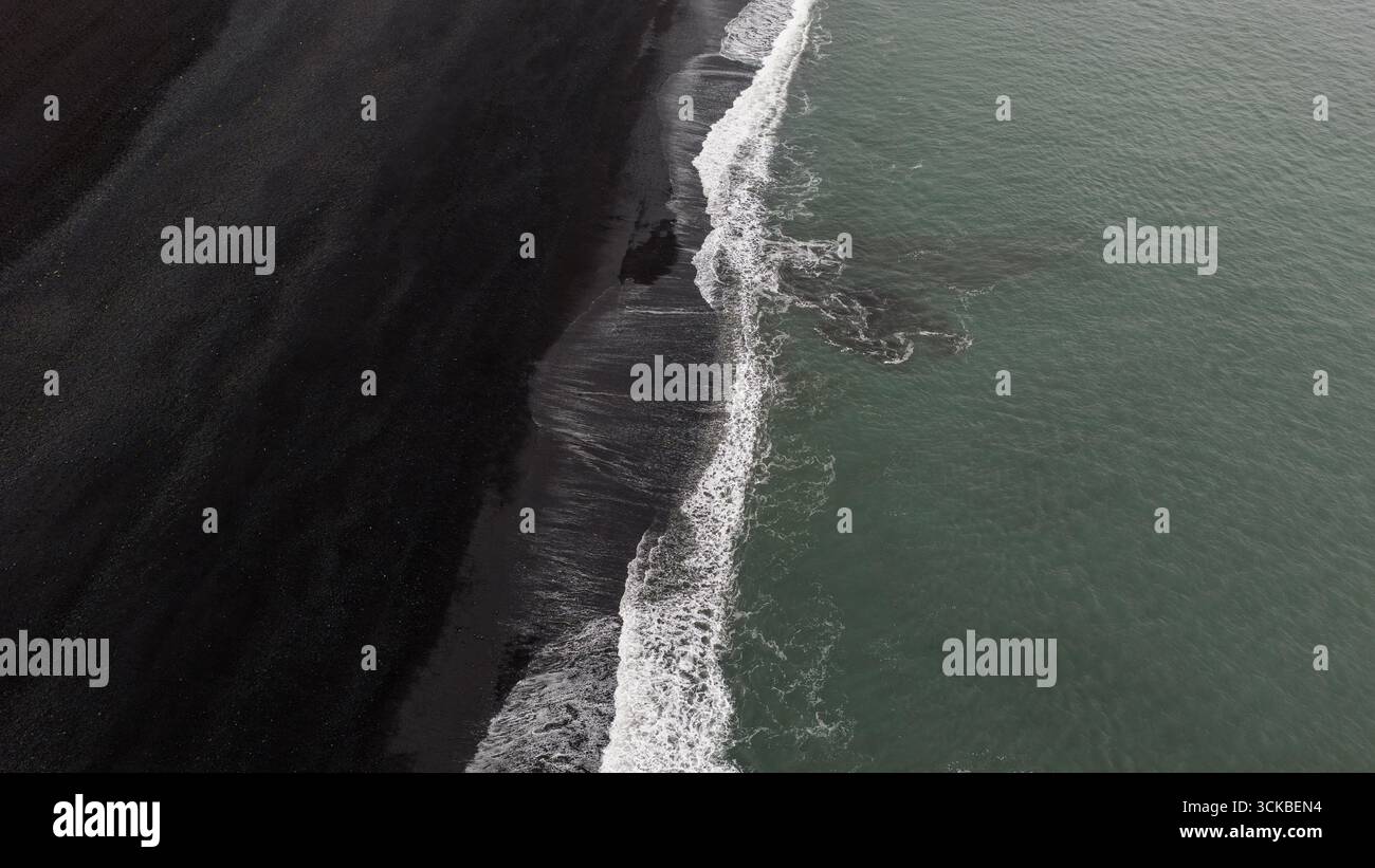 Aus der Vogelperspektive auf Islands vulkanische Küste mit schwarzem Sand trifft auf türkisfarbenes Meereswasser, mit scharfen Kontrasten und natürlichen Texturen. Stockfoto