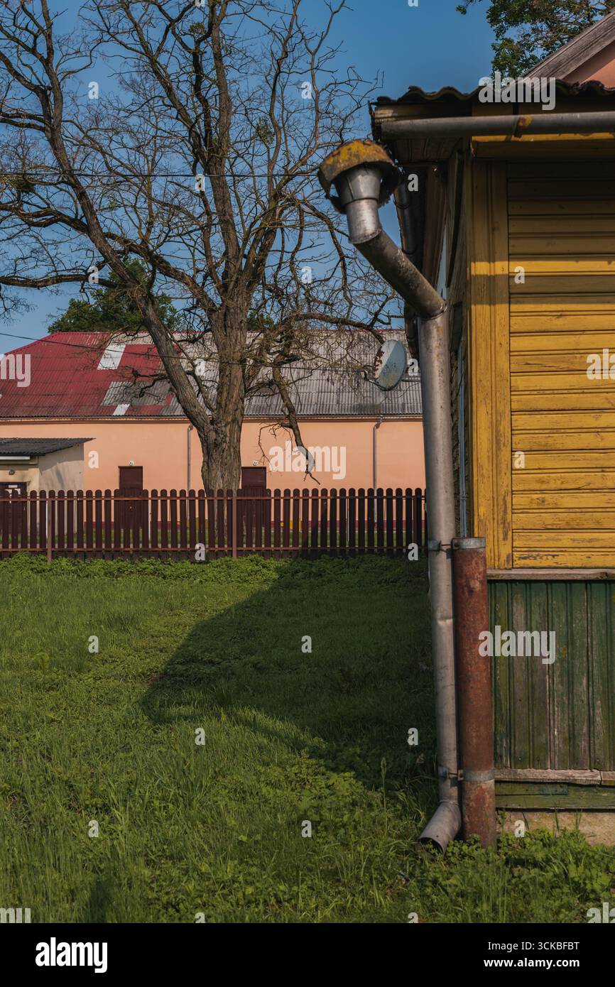 Ein gelbes Holzhaus mit grünen Akzenten und einem Metallabflussrohr steht neben einem Laubbaum, einem roten Gebäude und einem Holzzaun in einer grasbewachsenen Landschaft Stockfoto