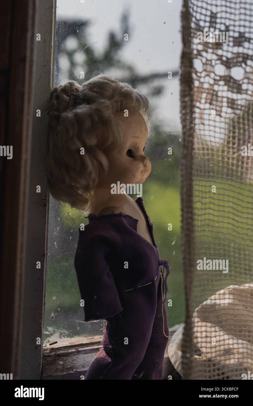 Eine alte Puppe mit lockigen blonden Haaren und einem violetten Outfit sitzt auf einer Fensterbank, mit Blick auf ein leicht schmutziges Fenster mit zerrissenen Spitzenvorhängen und Bäumen Stockfoto