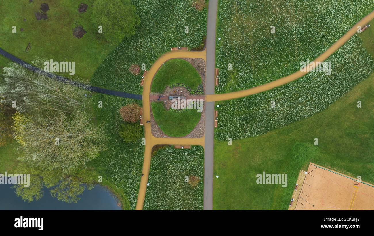 Blick aus der Vogelperspektive auf einen landschaftlich gestalteten Park mit sich kreuzenden Wegen, einem zentralen Rundgang, einem Teich, verstreuten Bäumen und einem sandigen Naherholungsgebiet. Stockfoto