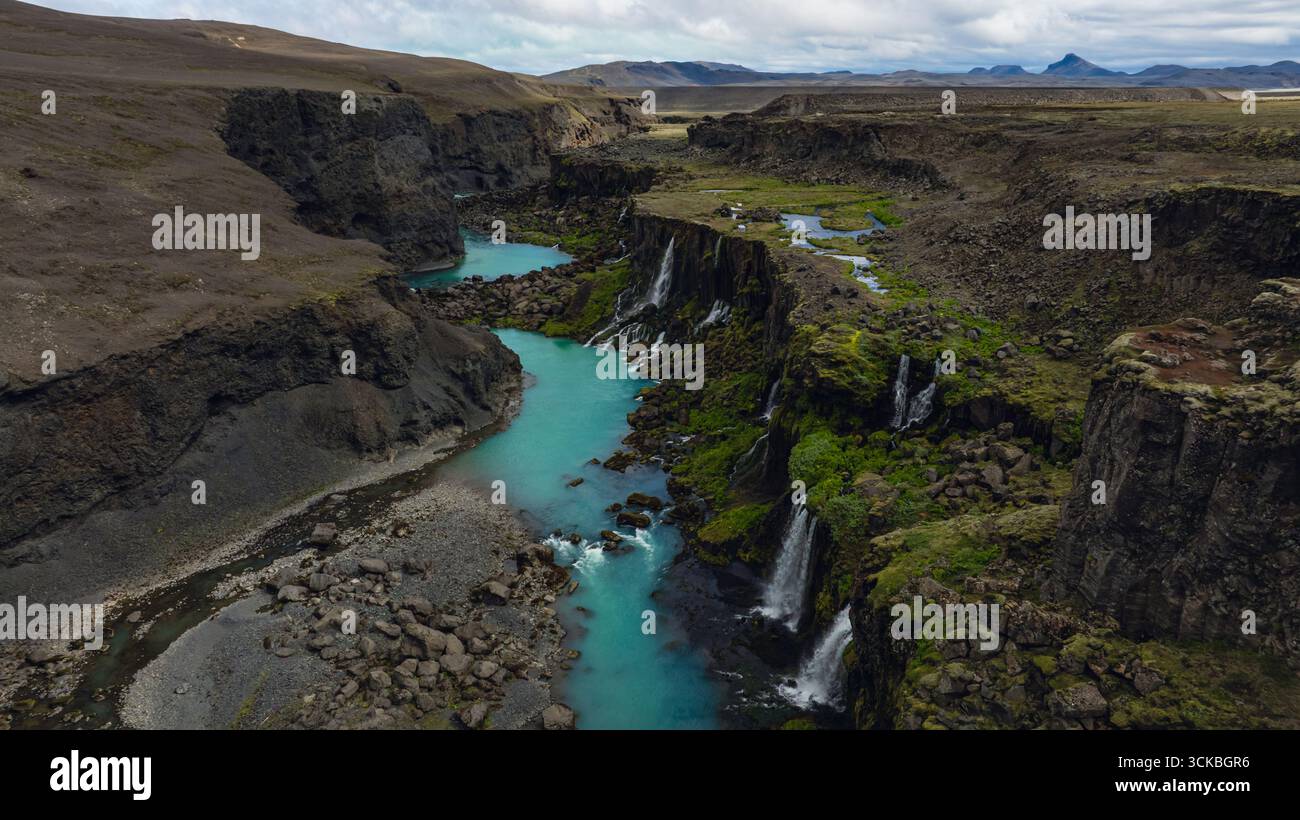 Dramatische Aussicht auf einen Canyon in Island mit einem türkisfarbenen Fluss, moosbedeckten Klippen mit Wasserfällen und fernen Bergen unter einem bewölkten Himmel Stockfoto