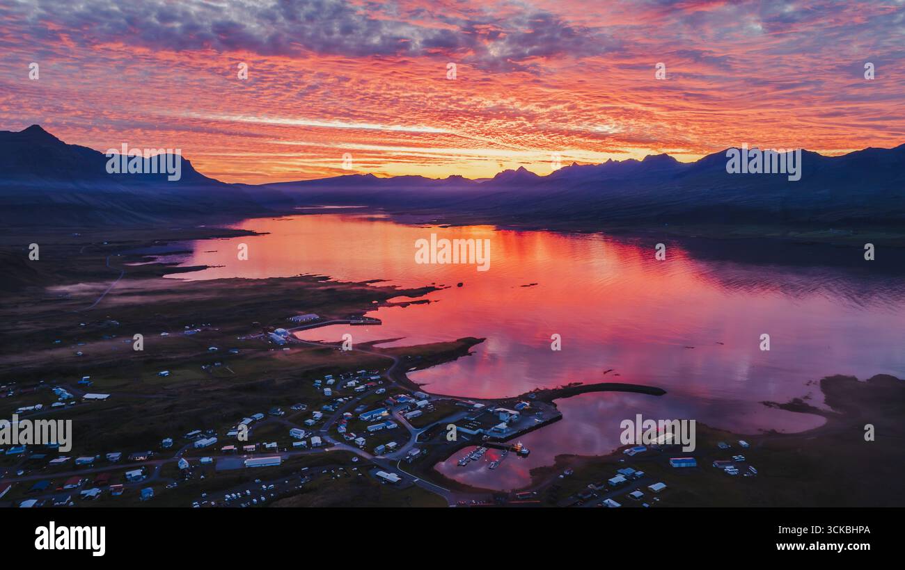 Eine kleine isländische Stadt liegt an einem ruhigen Fjord, umgeben von Bergen. Der Sonnenuntergangshimmel spiegelt sich auf dem Wasser und schafft eine ruhige und lebendige Szene. Stockfoto