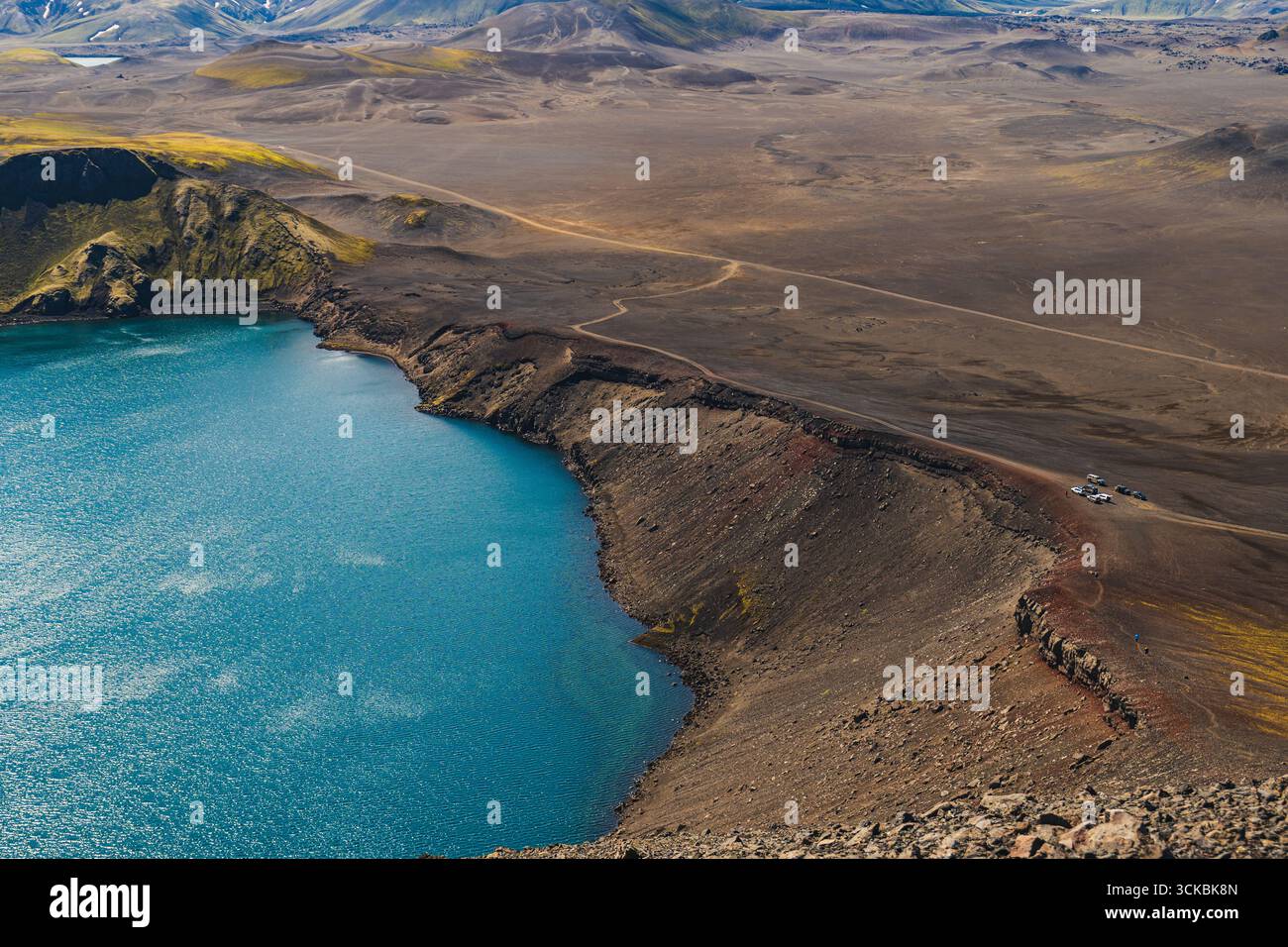 Ein lebendig blauer vulkanischer Kratersee, umgeben von zerklüftetem, kargem Gelände im isländischen Hochland, mit unbefestigten Straßen, karger Vegetation und weit entferntem Fahrzeug Stockfoto