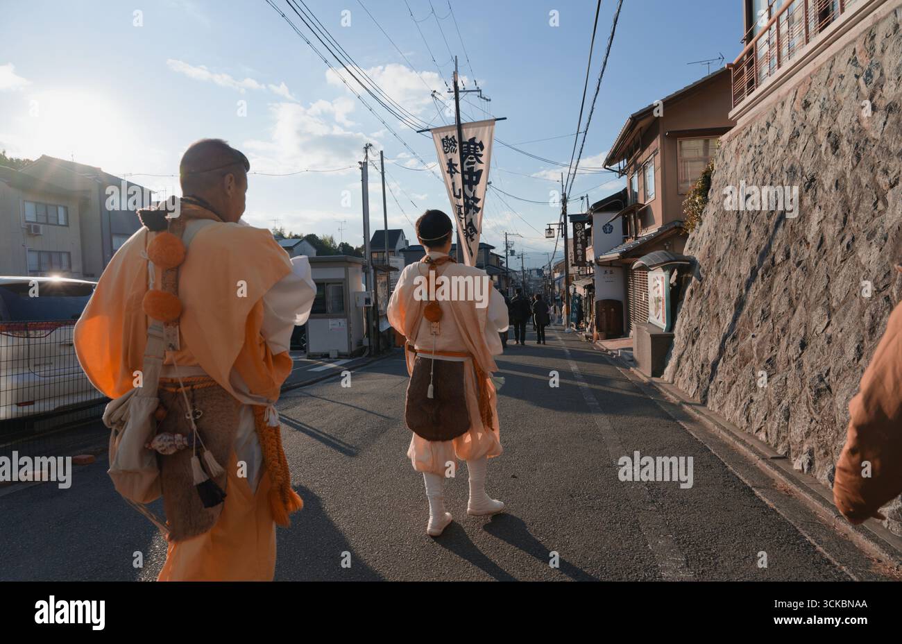 Mönche in Gewändern spazieren während des Tōka Ebisu Festivals entlang einer sonnigen Kyoto-Straße, tragen rituelle Banner, während sie lokale Geschäfte segnen Stockfoto
