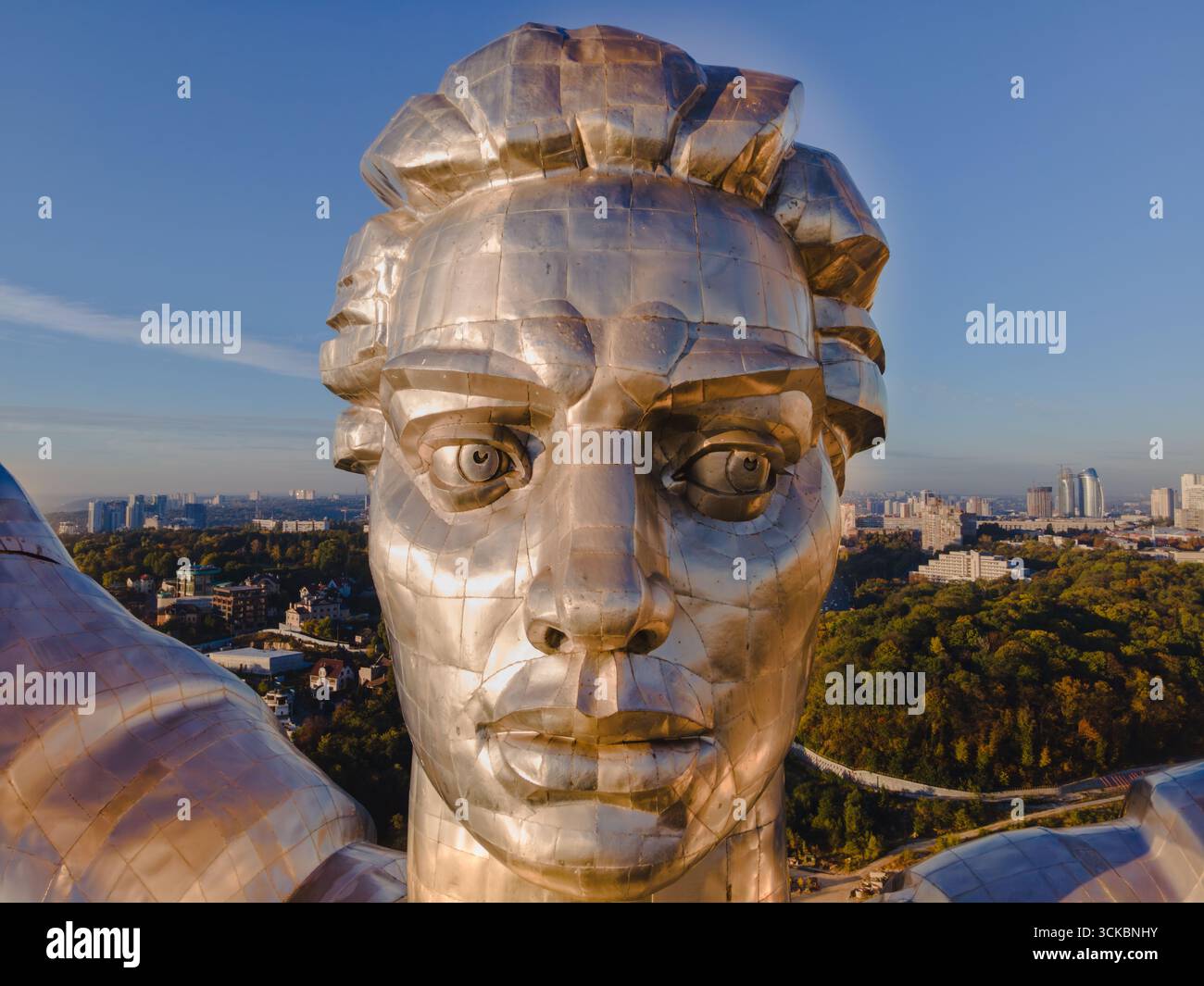 Nahaufnahme des Mutterlanddenkmals in Kiew, Ukraine, mit detaillierten Details, städtischen Gebäuden und Grün unter einem klaren blauen Himmel. Stockfoto