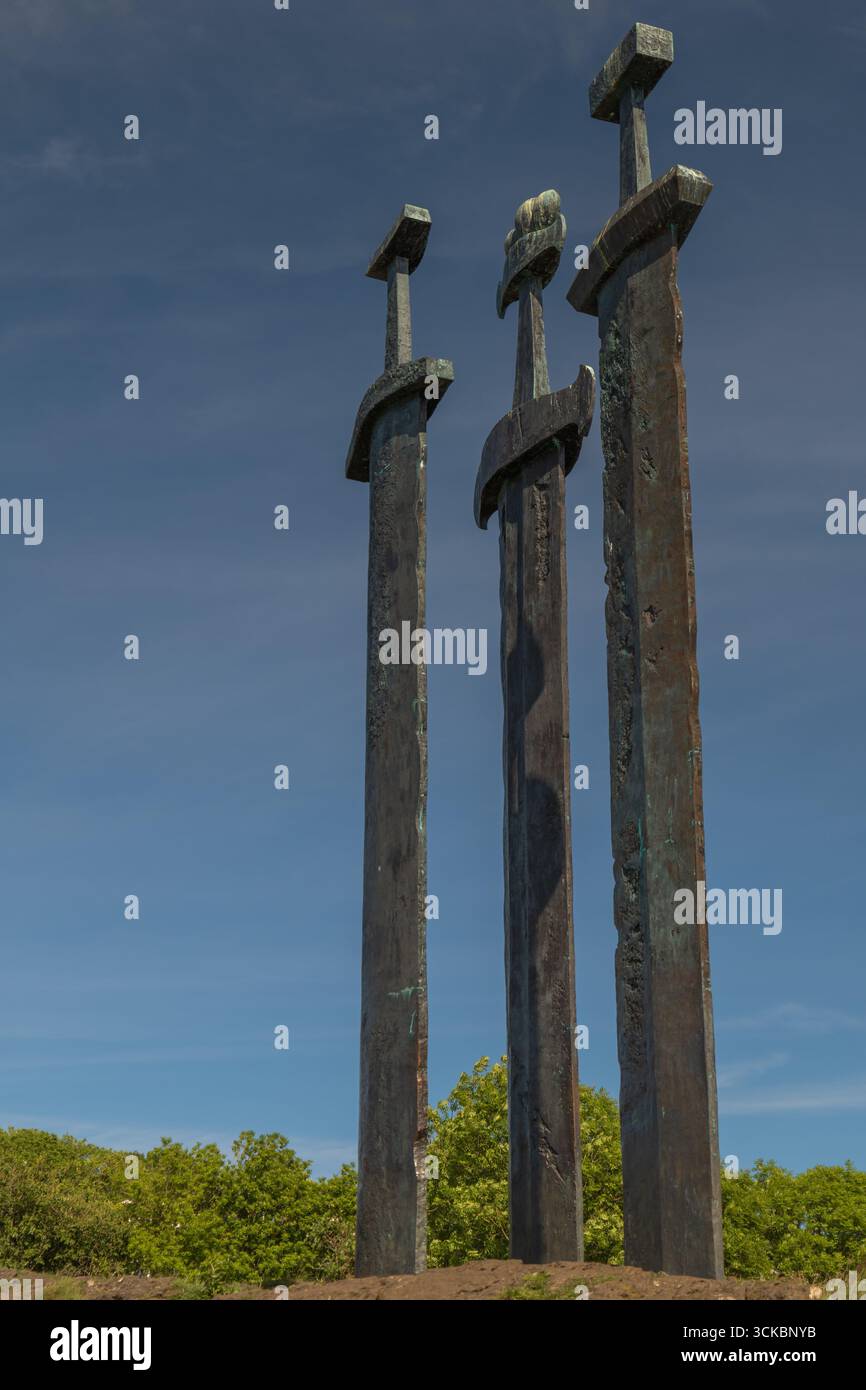 Das Sverd i Fjell Monument in Stavanger, Norwegen, zeigt drei große Bronzeschwerter, die in Felsen eingebettet sind und sich vor einem klaren blauen Himmel mit Grün darunter befinden Stockfoto