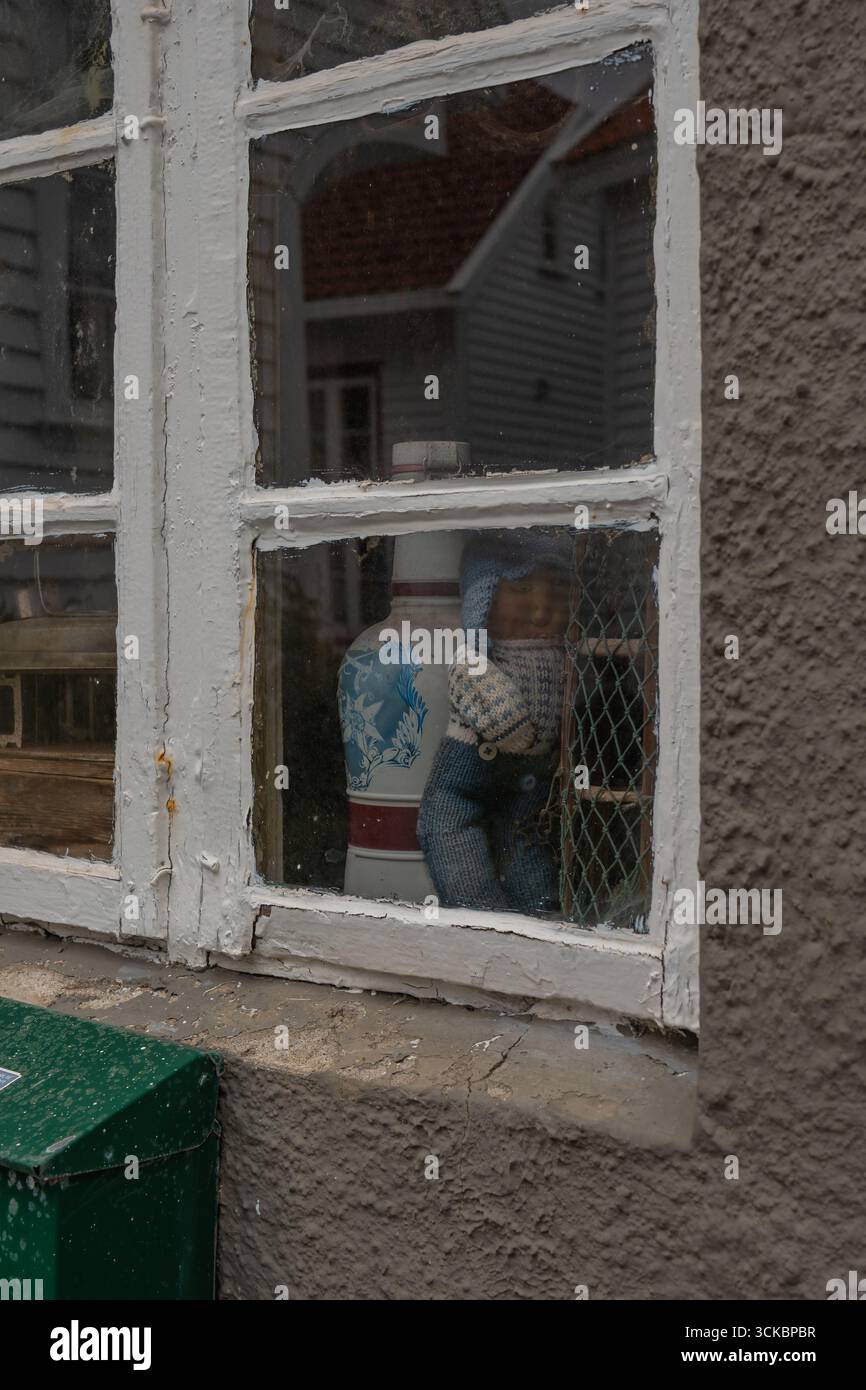 Nahaufnahme eines verwitterten, weiß gerahmten Fensters mit grünem Briefkasten, das Häuser im norwegischen Stil in einer Küstenstadt widerspiegelt. Stockfoto