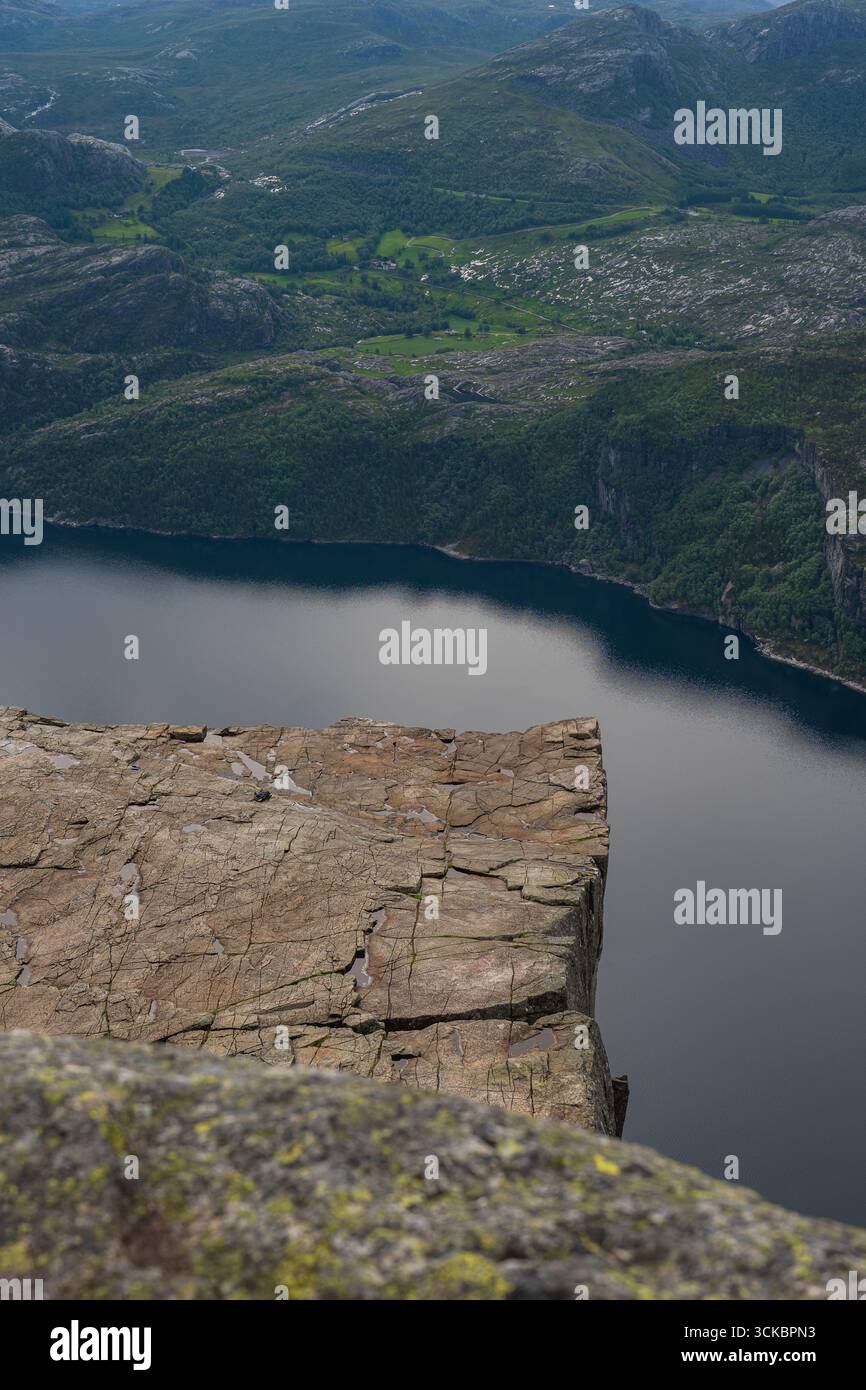 Blick auf Preikestolen's zerklüftete flache Spitze, mit dem ruhigen Wasser des Lysefjords und grünen Hügeln im Hintergrund, mit kleinen Siedlungen und natürlichen Scener Stockfoto