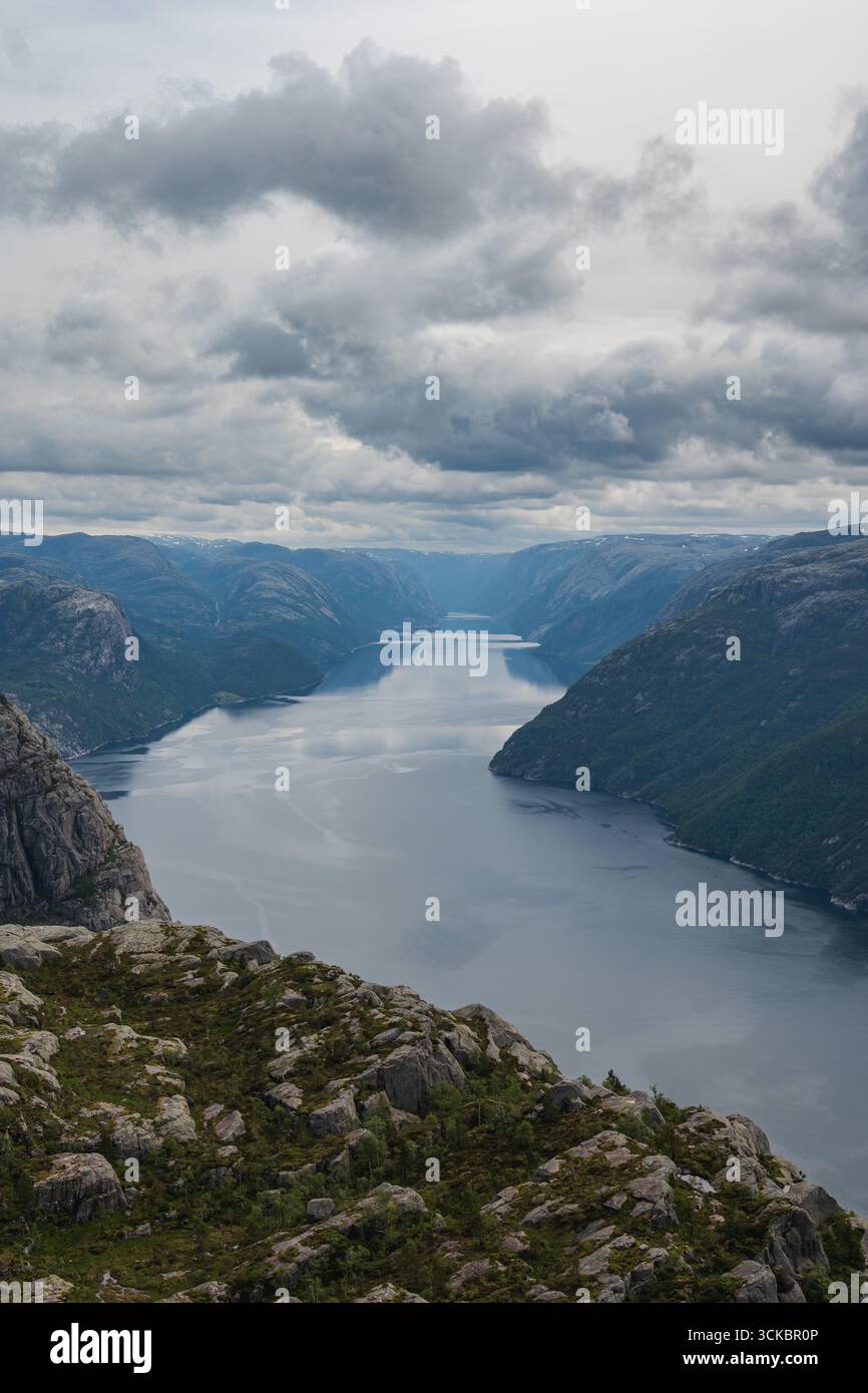 Das ruhige Wasser von Lysefjord erstreckt sich bis in die Ferne, umgeben von zerklüfteten Klippen und üppigem Grün unter einem dramatischen, wolkengefüllten Himmel. Stockfoto