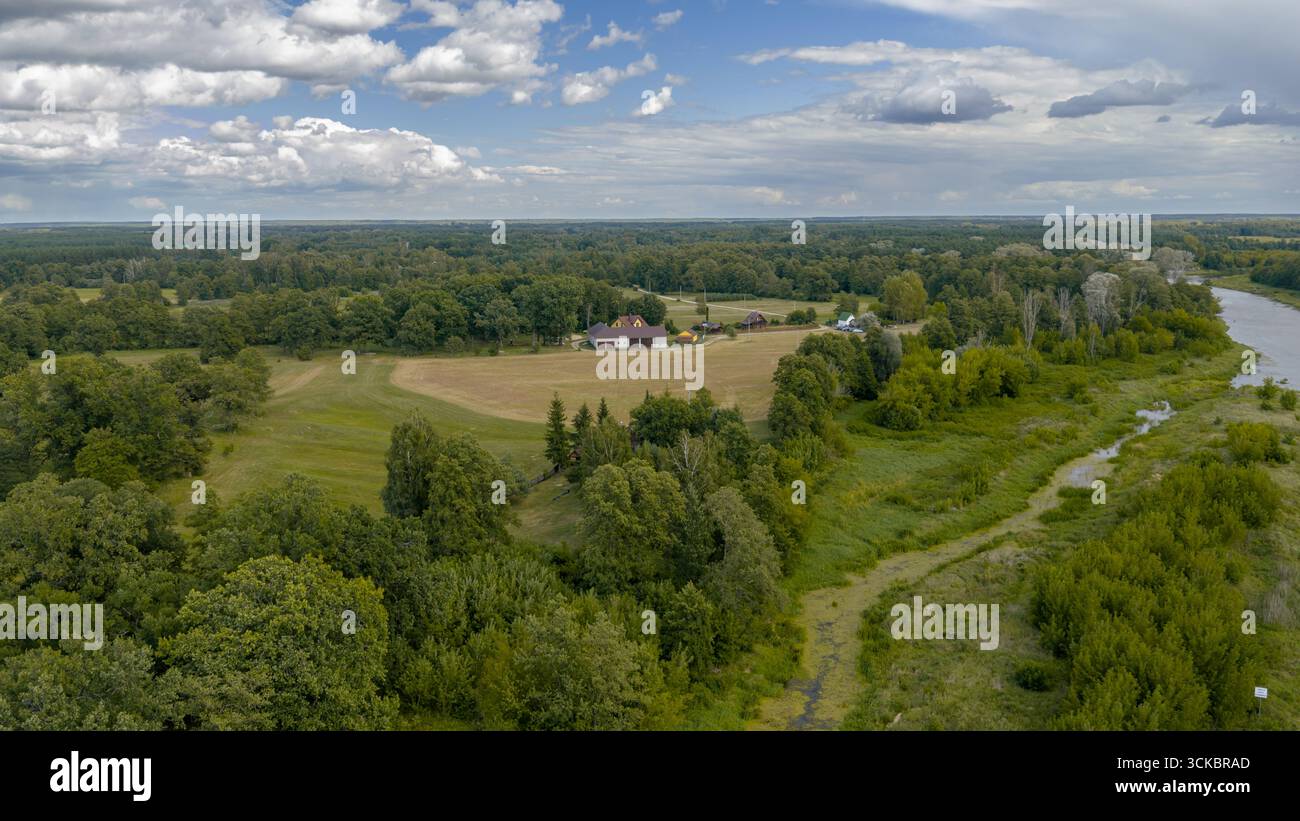 Ein sich windender Fluss fließt durch Nowy Lubiel in Polen, umgeben von Grün und Wald. Felder und eine Gruppe von Gebäuden, darunter ein rotes überdachtes Haus, A Stockfoto