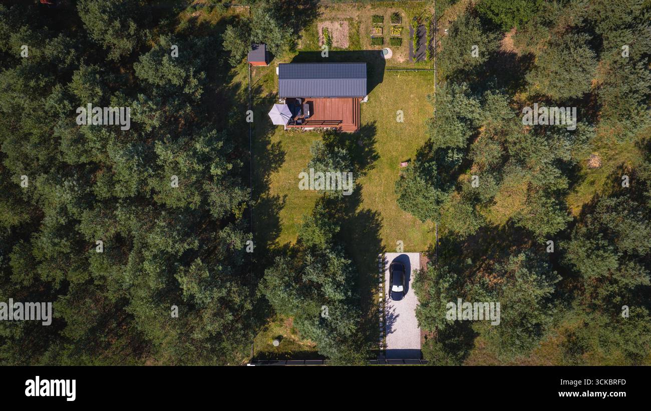 Luftbild eines modernen Hauses mit dunklem Dach, Holzdeck, Rasen, Garten und Auffahrt, umgeben von dichtem Wald in der Nähe von Nowy Lubiel, Polen. Stockfoto