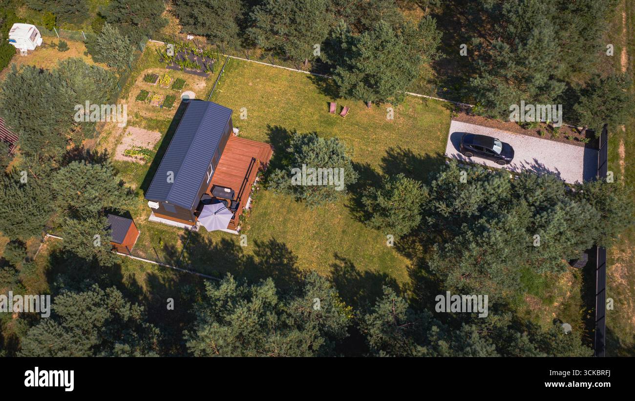 Luftbild eines kleinen Hauses mit dunklem Dach, Holzdeck, Garten, Schottereinfahrt und schwarzem Auto, in einem Wald in der Nähe von Nowy Lubiel, Polen. Stockfoto