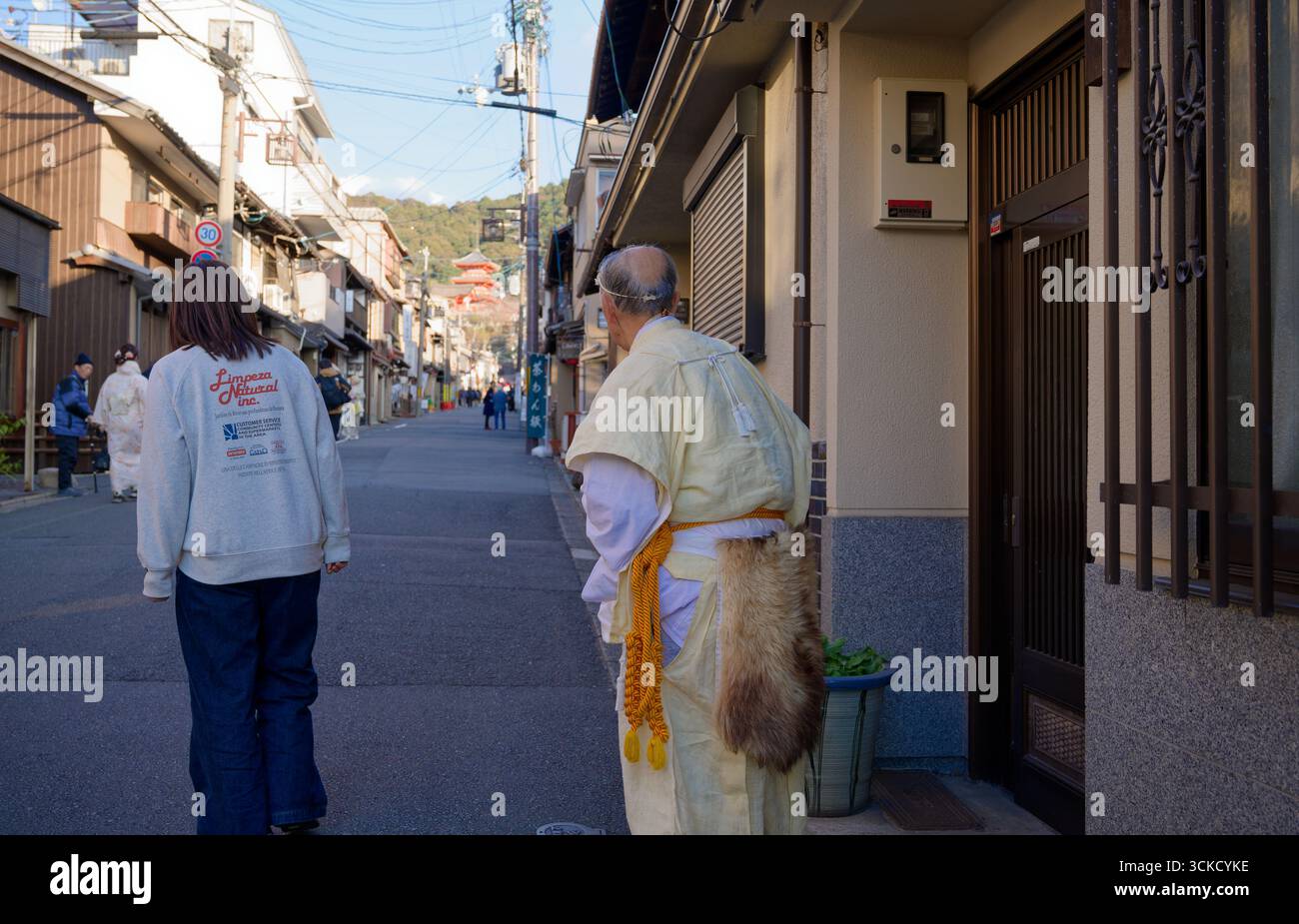 Ein älterer Mönch in gelben Gewändern mit einem Pelzgürtel hält während des Tōka Ebisu Festivals in einer ruhigen Straße in Kyoto inne Stockfoto