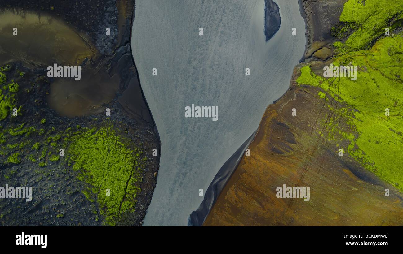 Luftbild eines silbrigen grauen Flusses, der durch Islands Landschaft schneidet, mit grünem Moos auf vulkanischem Boden und kontrastierenden Braun- und Orangetönen. Stockfoto