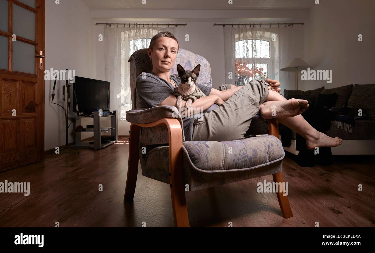 Eine Frau mittleren Alters mit grauen kurzen Haaren hält ihre kanadische Sphynx-Katze in den Armen und lächelt sanft in einem gut beleuchteten Wohnzimmer mit Holzböden und so Stockfoto