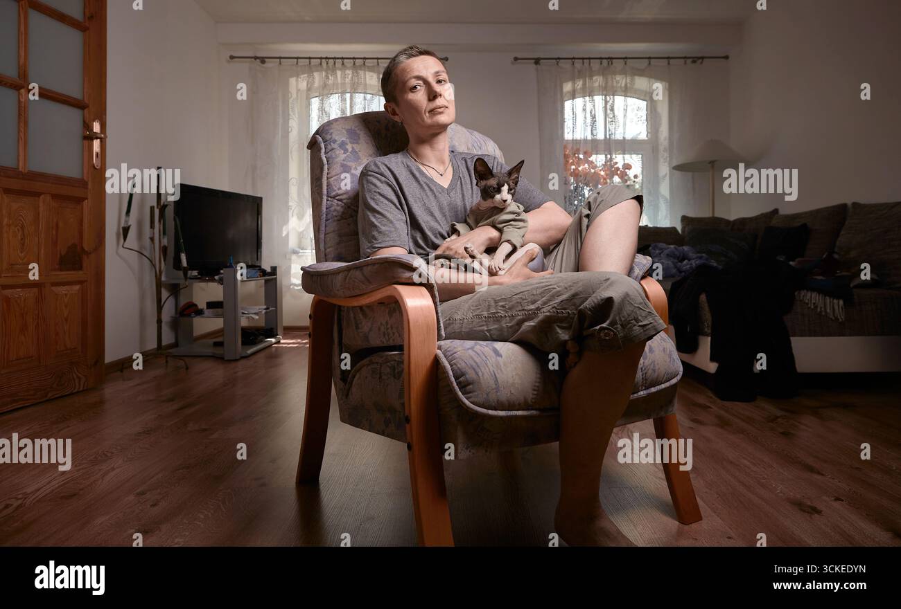 Eine Frau mittleren Alters mit grauen, kurzen Haaren sitzt bequem mit ihrer kanadischen Sphynx-Katze auf ihrem Schoß in einem modernen Wohnzimmer. Sie verkörpert Komfort, Frieden, A Stockfoto