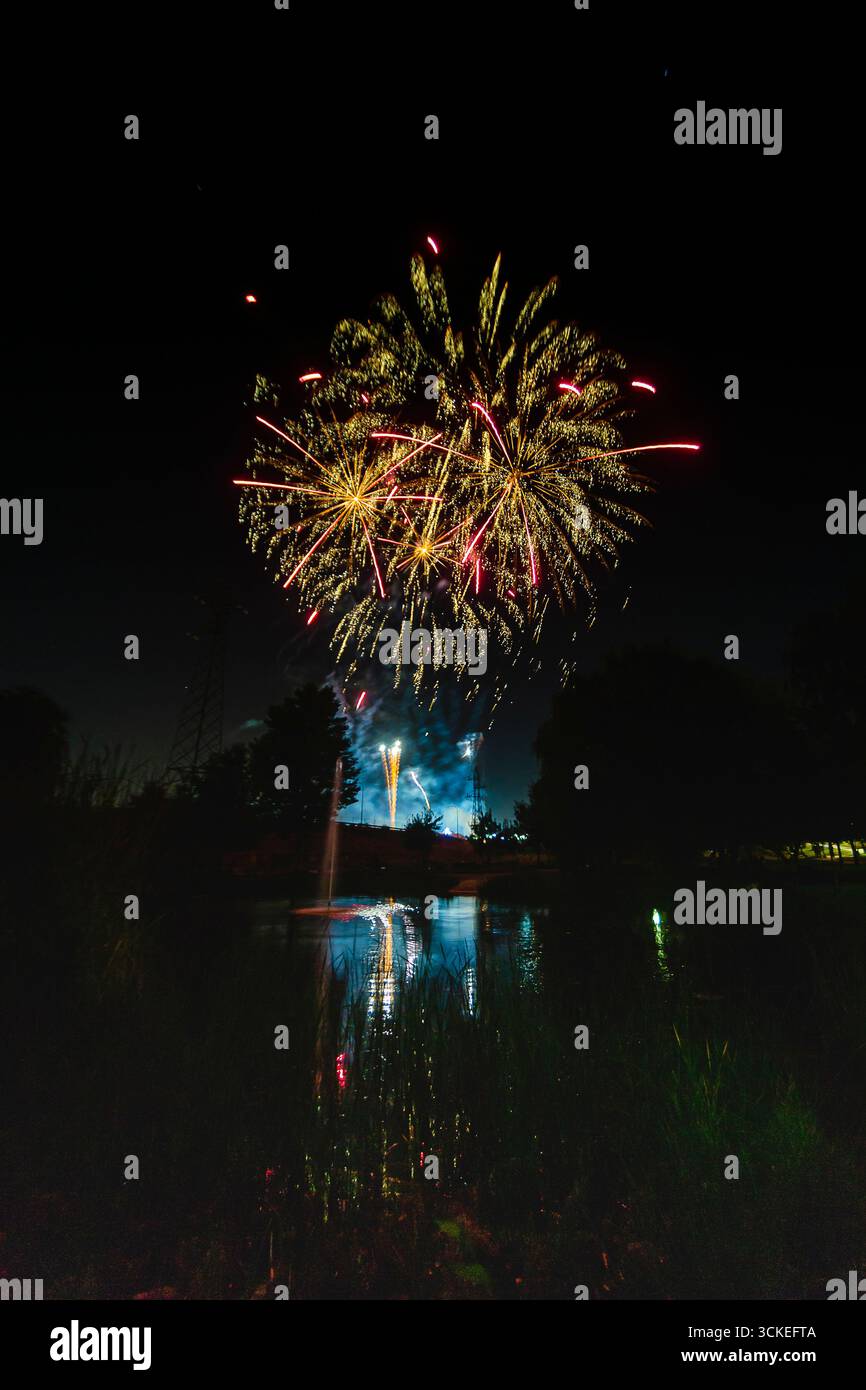 Ein helles Feuerwerk erleuchtet den Nachthimmel über einem ruhigen See und reflektiert bunte Schauplätze im Wasser. Stockfoto