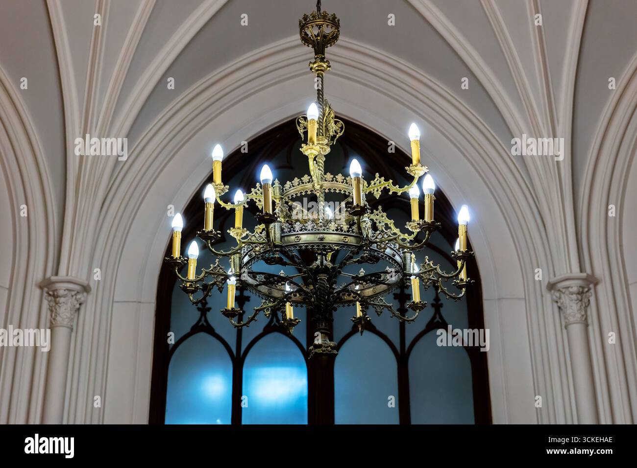 Großer Kronleuchter aus Metall mit Kerzenlampen, die unter gotischen Rippengewölben über Buntglasfenstern aufgehängt sind. Stimmungsvolle sakrale Architektur su Stockfoto