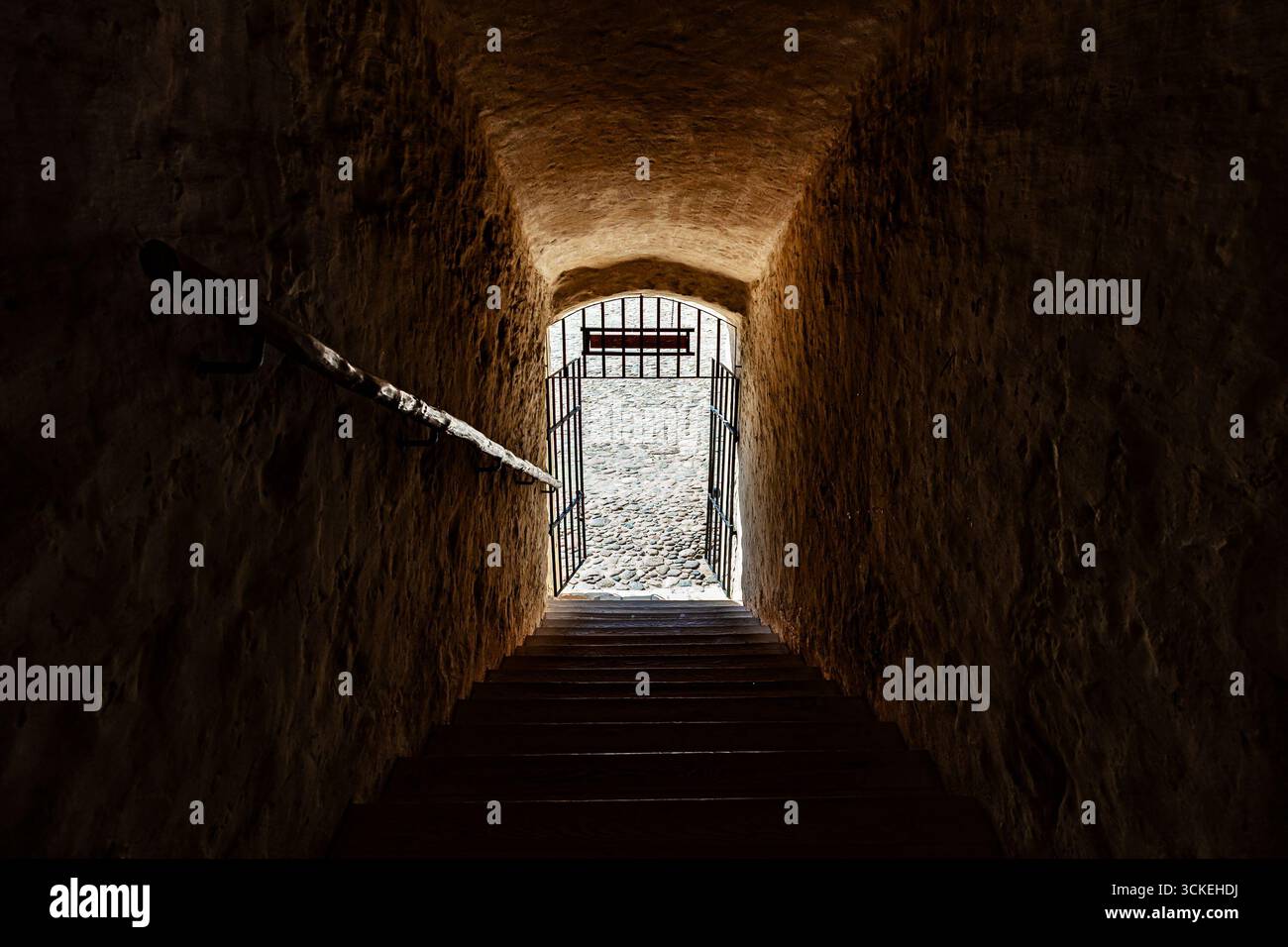 Ein schmaler Tunnel aus rauem Stein führt zu einem sonnendurchfluteten schmiedeeisernen Tor, das dramatische Kontraste und Geheimnisse schafft. Erinnert an Geschichte, Sicherheit und Reise Stockfoto