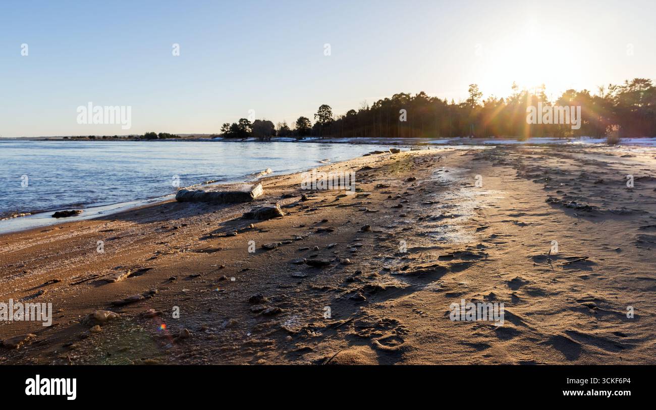 Eine ruhige Winterküstenlandschaft im Morgenlicht mit gefrorenem Sandstrand, ruhigem Wasser und strahlendem Sonnenaufgang. Die ruhige sce Stockfoto