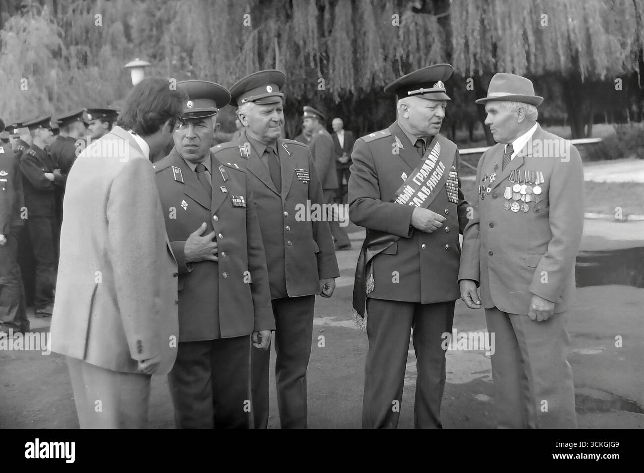 Ein klassisches „Treffen der Generationen“ in Sloviansk in den 1970er Jahren, wo eine Gruppe von angesehenen Veteranen des Großen Vaterländischen Krieges, die mit Orden und Medaillen geschmückt sind und zu denen auch ein Ehrenbürger der Stadt gehört, während einer Feier zum Siegtag Gespräche mit einem jüngeren Mann führt. Dieses Foto ist zutiefst symbolisch für die Kontinuität und die Übertragung des historischen Gedächtnisses, ein ideologisches Schlüsselkonzept, das in der Sowjetgesellschaft während der Breschnew-Ära gefördert wurde, um die heroische Vergangenheit mit der heutigen Generation zu verbinden Stockfoto