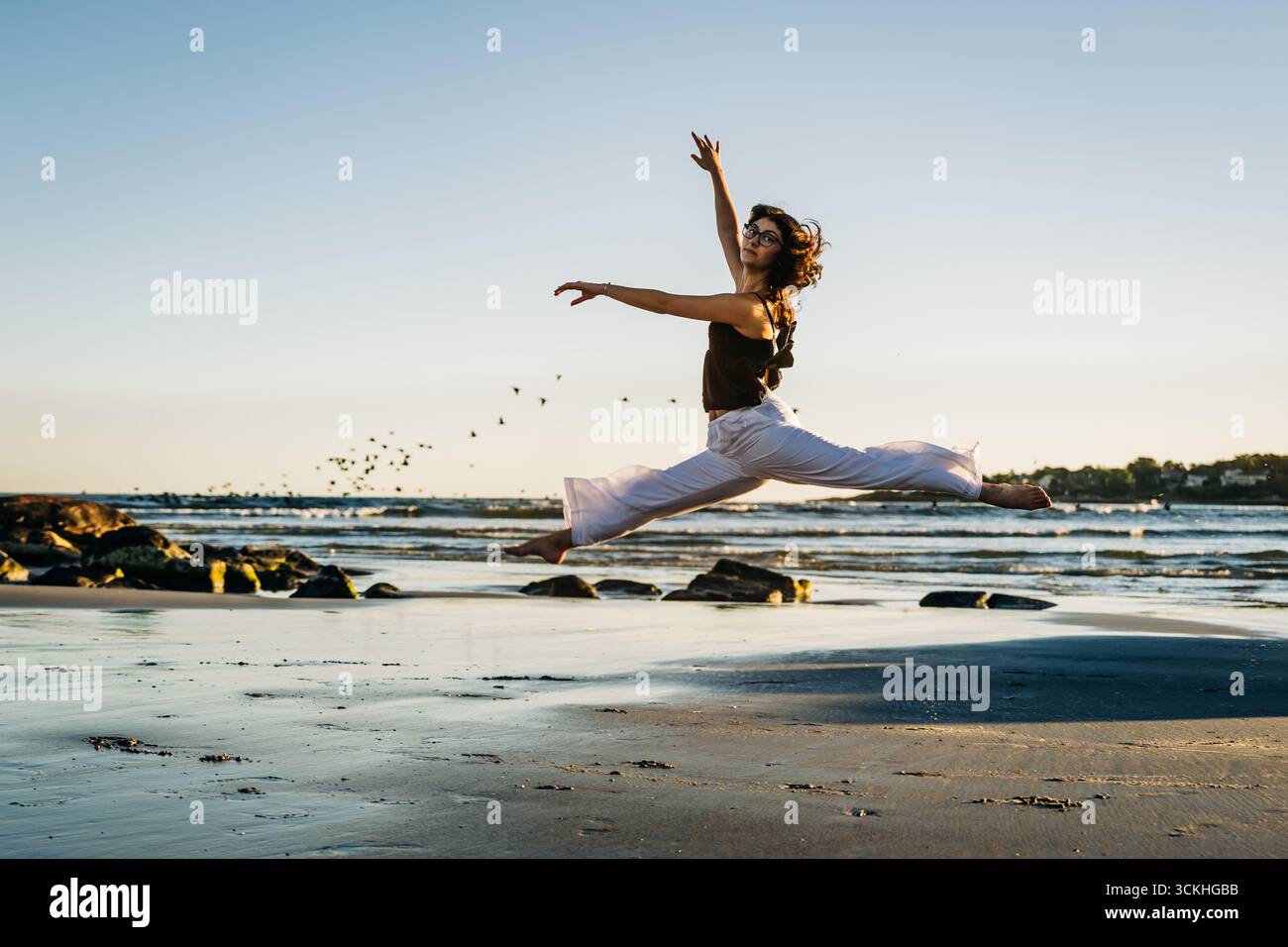 Junge Ballerina, die Splits aufführt, springen mit Vögeln am Strand Stockfoto
