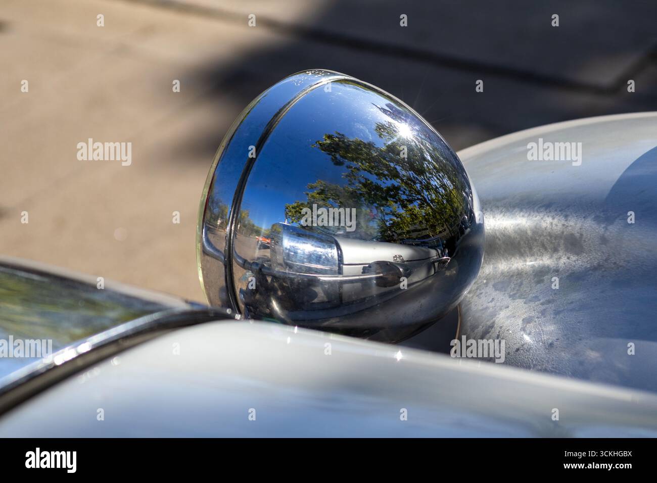 Glänzender Chrom-Scheinwerfer, der die Reflexionen umliegender Bäume und den leuchtenden blauen Himmel in einem sorgfältig restaurierten Oldtimer einfängt Stockfoto