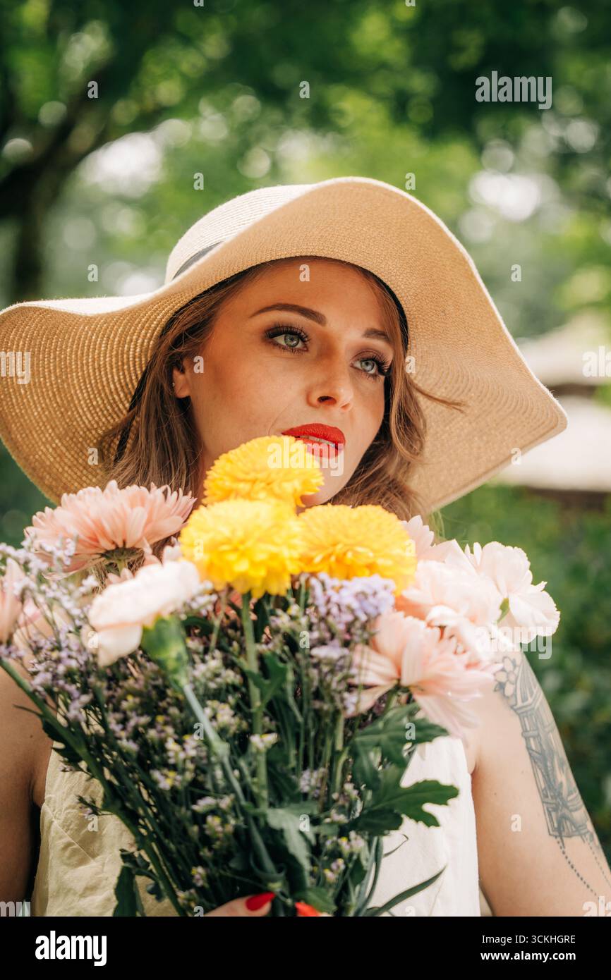 Stilvolle Frau mit Blumenstrauß und Sonnenhut im Park Stockfoto