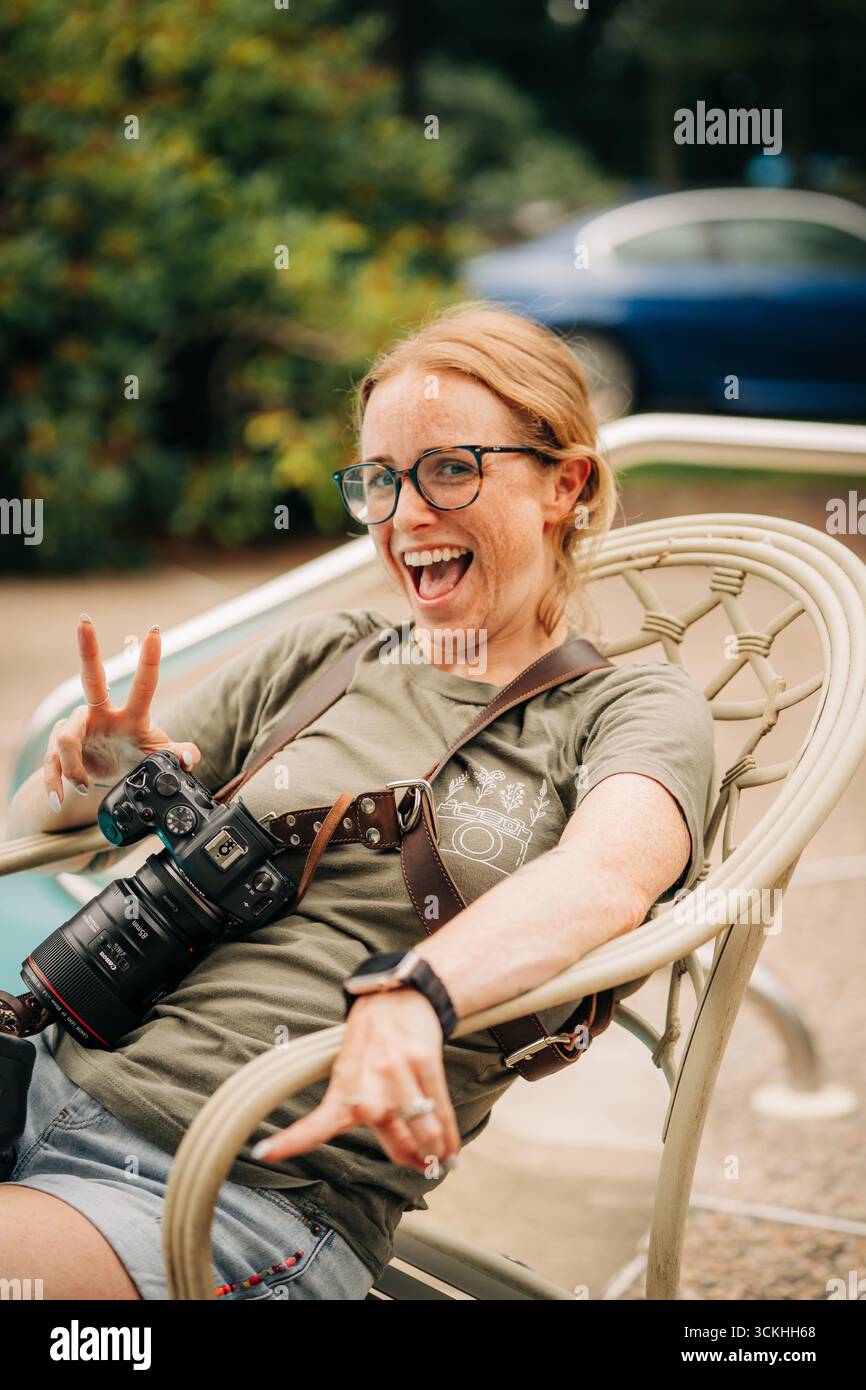 Lächelnde Fotografin, die ein Friedensschild blitzt, während sie draußen sitzt Stockfoto