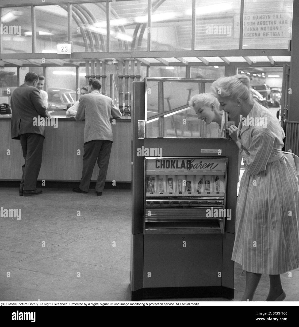 Verkaufsautomat 1957. Eine blonde Frau lehnt sich an den Automaten, der Schokoladentafeln verschiedener Art anbietet. Der typische, 1957 für Frauen beliebte Rock war ein breiter, ausgestellter A-Linie oder Kreisrock, der oft mit einem Gürtel an der Taille verschlungen wurde, um eine Sanduhr-Silhouette zu erzeugen. Es besteht aus Stoffen wie Baumwolle, Tüll oder Chiffon, fällt normalerweise auf mittlere Wadenlänge und wurde mit Unterröcken getragen, um mehr Volumen zu erhalten. Der Stil war lebendig, feminin und ikonisch mit der Mode des „New Look“ der 1950er Jahre. Conard Ref. 3496 Stockfoto