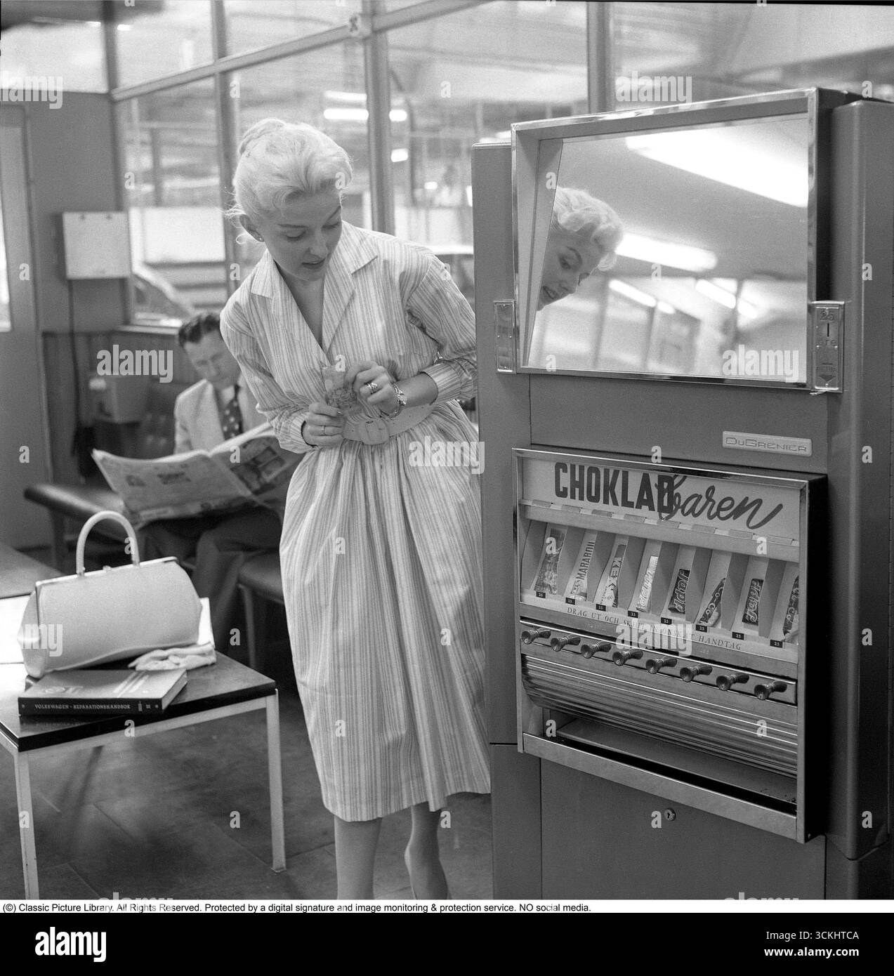 Verkaufsautomat 1957. Eine blonde Frau lehnt sich an den Automaten, der Schokoladentafeln verschiedener Art anbietet. Der typische, 1957 für Frauen beliebte Rock war ein breiter, ausgestellter A-Linie oder Kreisrock, der oft mit einem Gürtel an der Taille verschlungen wurde, um eine Sanduhr-Silhouette zu erzeugen. Es besteht aus Stoffen wie Baumwolle, Tüll oder Chiffon, fällt normalerweise auf mittlere Wadenlänge und wurde mit Unterröcken getragen, um mehr Volumen zu erhalten. Der Stil war lebendig, feminin und ikonisch mit der Mode des „New Look“ der 1950er Jahre. Conard Ref. 3496 Stockfoto