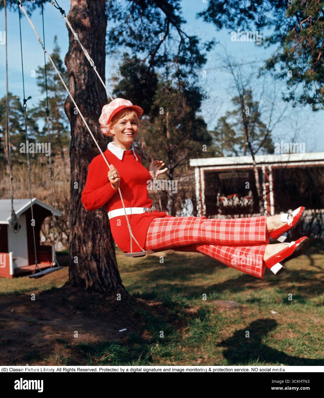 Mode der 1970er Jahre Die schwedische Schauspielerin Christina Schollin trägt die typische Mode der 70er Jahre. Ein roter Pullover und eine Hose mit passenden Schuhen und Mützen. Ein farbenfrohes Outfit, wenn man in einer Schaukel auf dem Foto im Garten ihres Hauses sitzt. Stockfoto