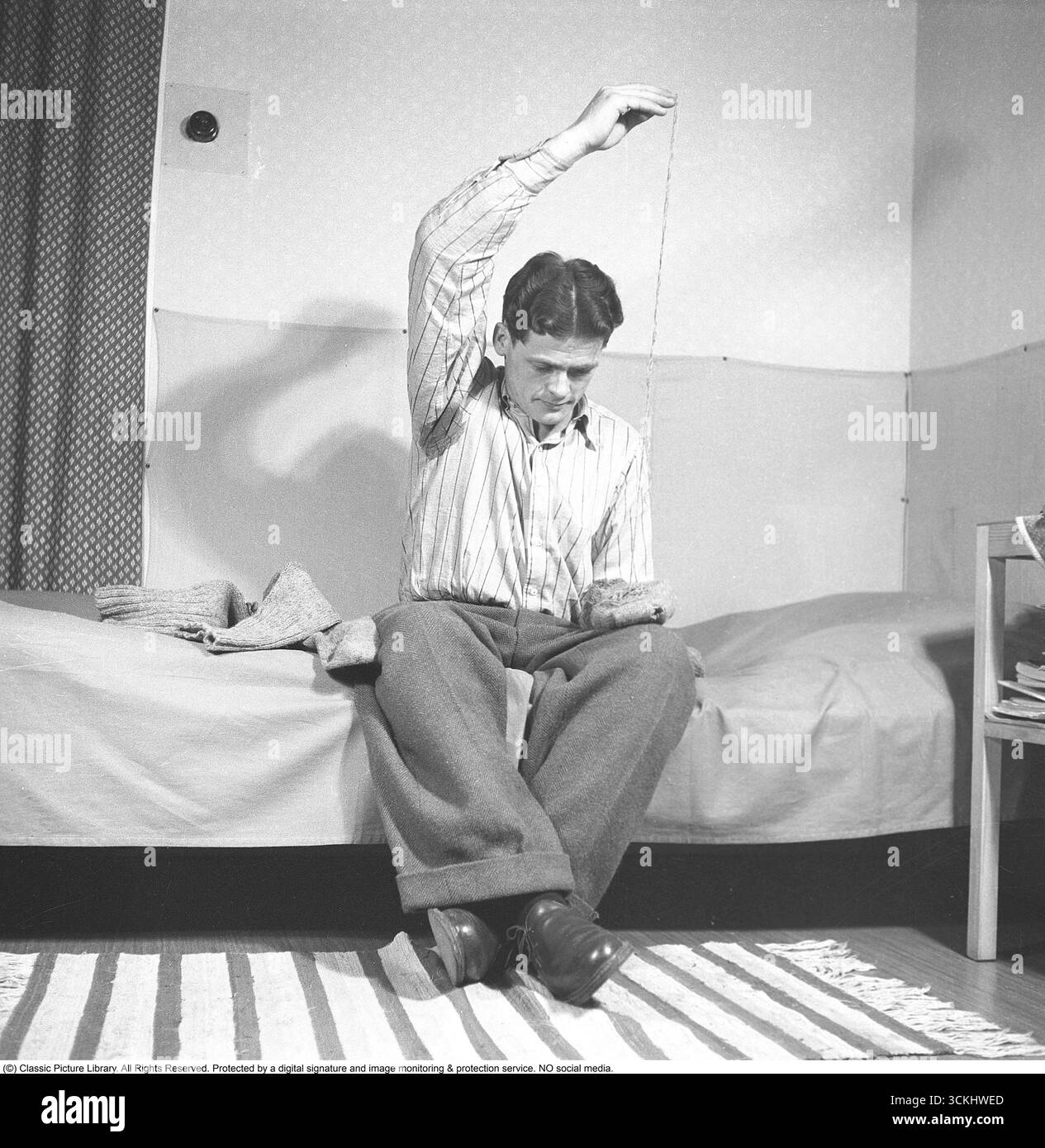 Am besten repariert man das Loch in der Socke... Ein Mann sitzt 1947 und repariert die Löcher in seinen Wollstrümpfen. Diese Szene spiegelt den Einfallsreichtum und die Sparsamkeit wider, die in der Nachkriegszeit üblich waren. 1947 stand Europa nach dem Zweiten Weltkrieg vor wirtschaftlichen Herausforderungen, was zu einer Kultur des Ausbesserns und Wiederverwendens von Gegenständen wie Kleidung führte. Diese Praxis war weit verbreitet, da die Menschen an Engpässe und Rationierungen angepasst wurden. (Kristoffersson, Ref. AF13-6) Stockfoto