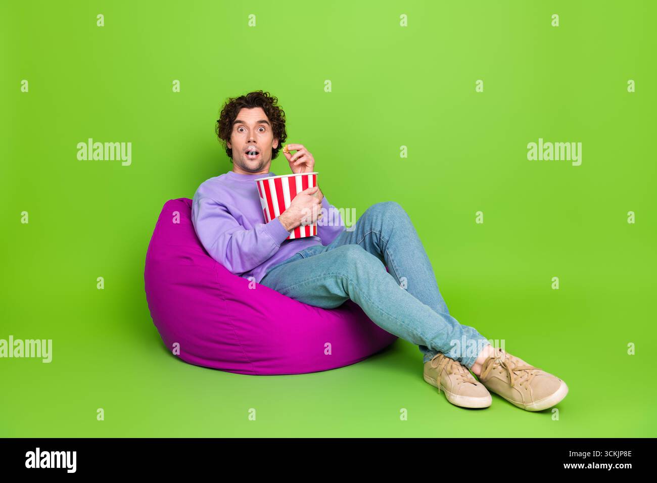 Junger Mann mit lockigem Haar, der einen Snack auf einem lila Sitzsack-Stuhl vor einem hellgrünen Hintergrund genießt Stockfoto