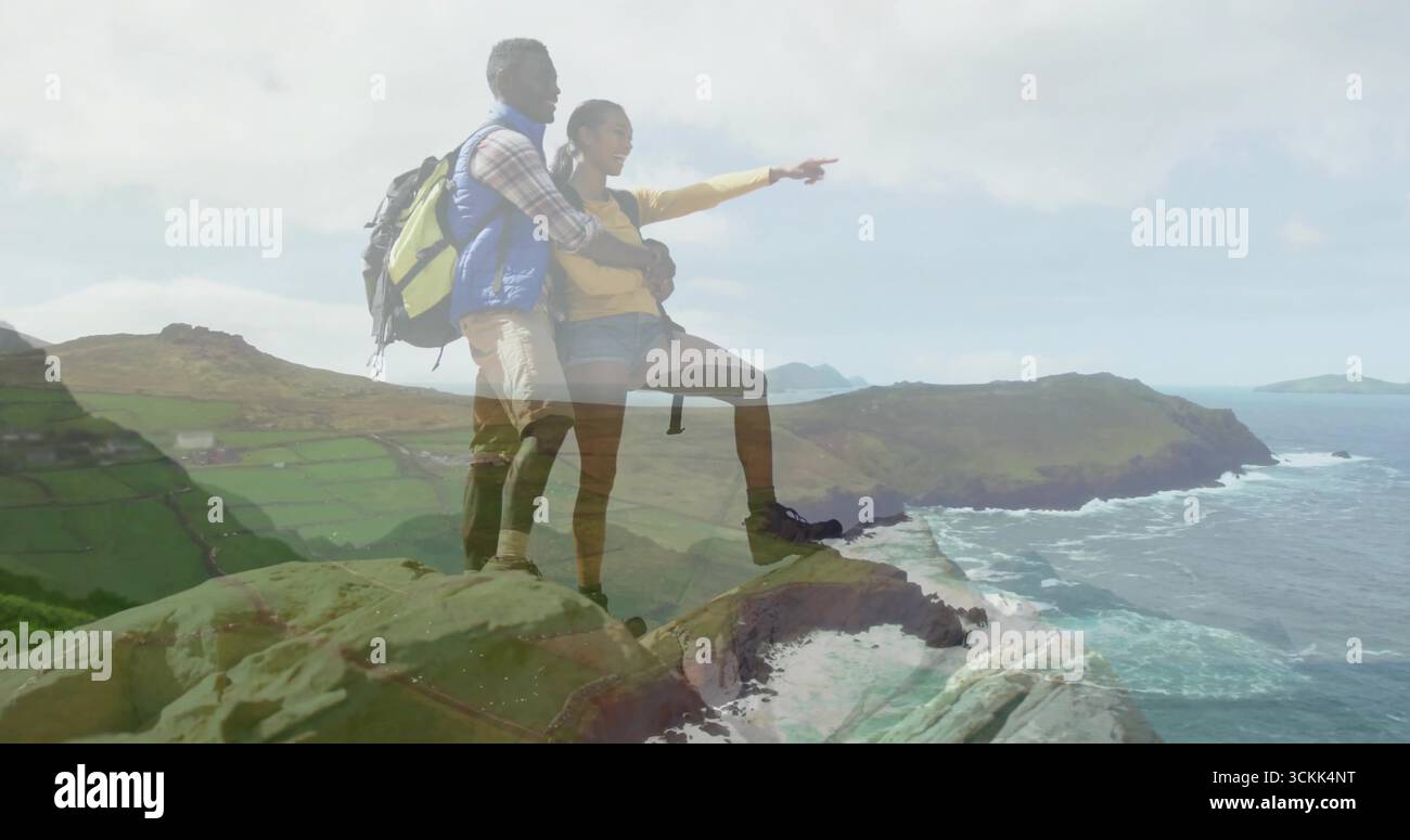 Ein schiefes Paar in gelber Windjacke, das von der Klippe aus auf das Meer zeigt, mit Wanderrucksack Stockfoto