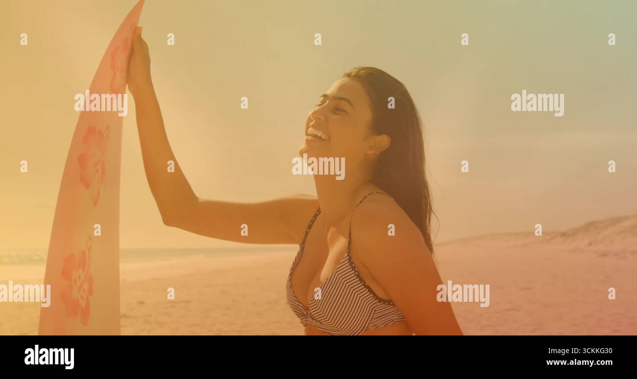 Lächelnde asiatische Frau in gestreiftem Bikini mit rosafarbenem Surfbrett am Strand bei Sonnenuntergang, Kopierraum Stockfoto