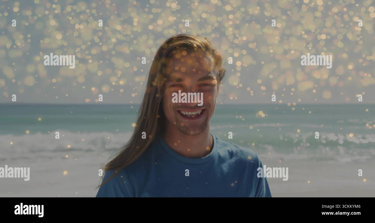 Lächelnder Mann mit blauem T-Shirt, der am ruhigen türkisfarbenen Meer am Strand steht, mit goldenen Bokeh-Überzügen Stockfoto