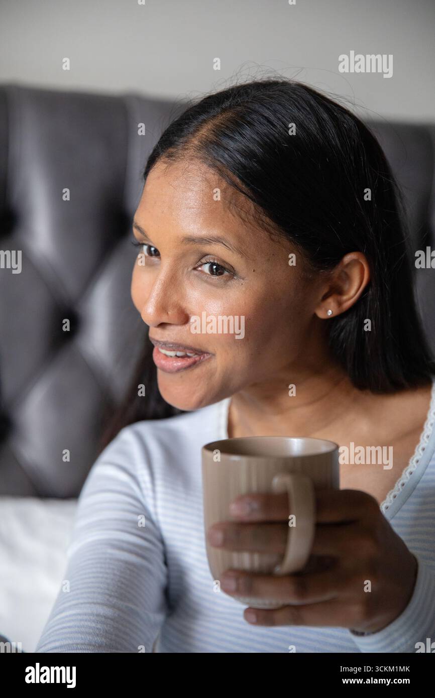 Indische Frau, die ein leichtes langärmeliges Oberteil trägt und einen Keramikbecher hält, der zu Hause auf dem Bett spricht Stockfoto