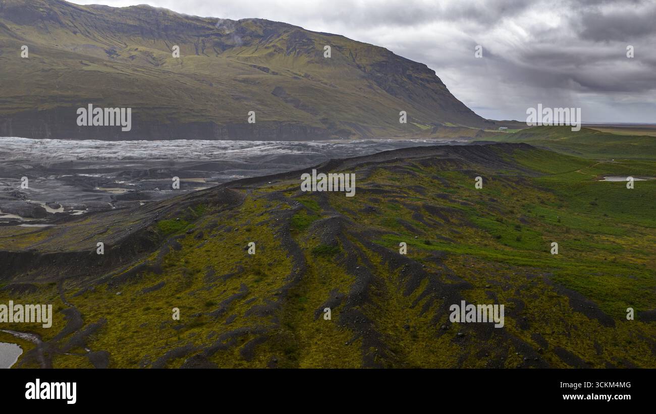 Zerklüftetes vulkanisches Gelände mit grünem Moos, einem Gletscher in der Mitte und einem gewaltigen Berg unter einem bedeckten Himmel in Islands dramatischer Landschaft. Stockfoto