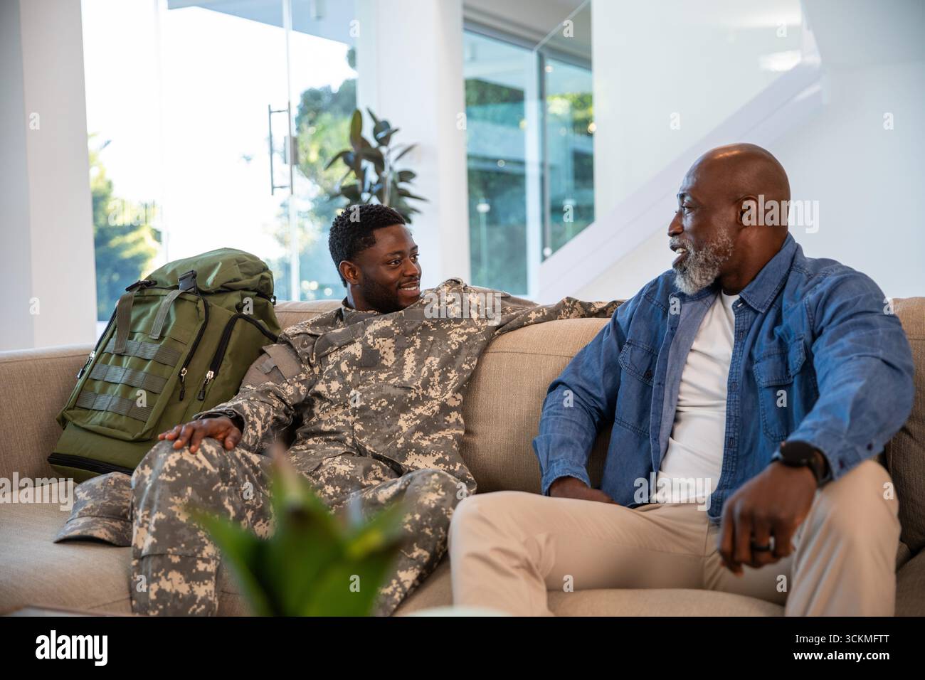 afroamerikaner Vater und Sohn sitzen zu Hause in Militäruniform mit Rucksack auf dem Sofa Stockfoto