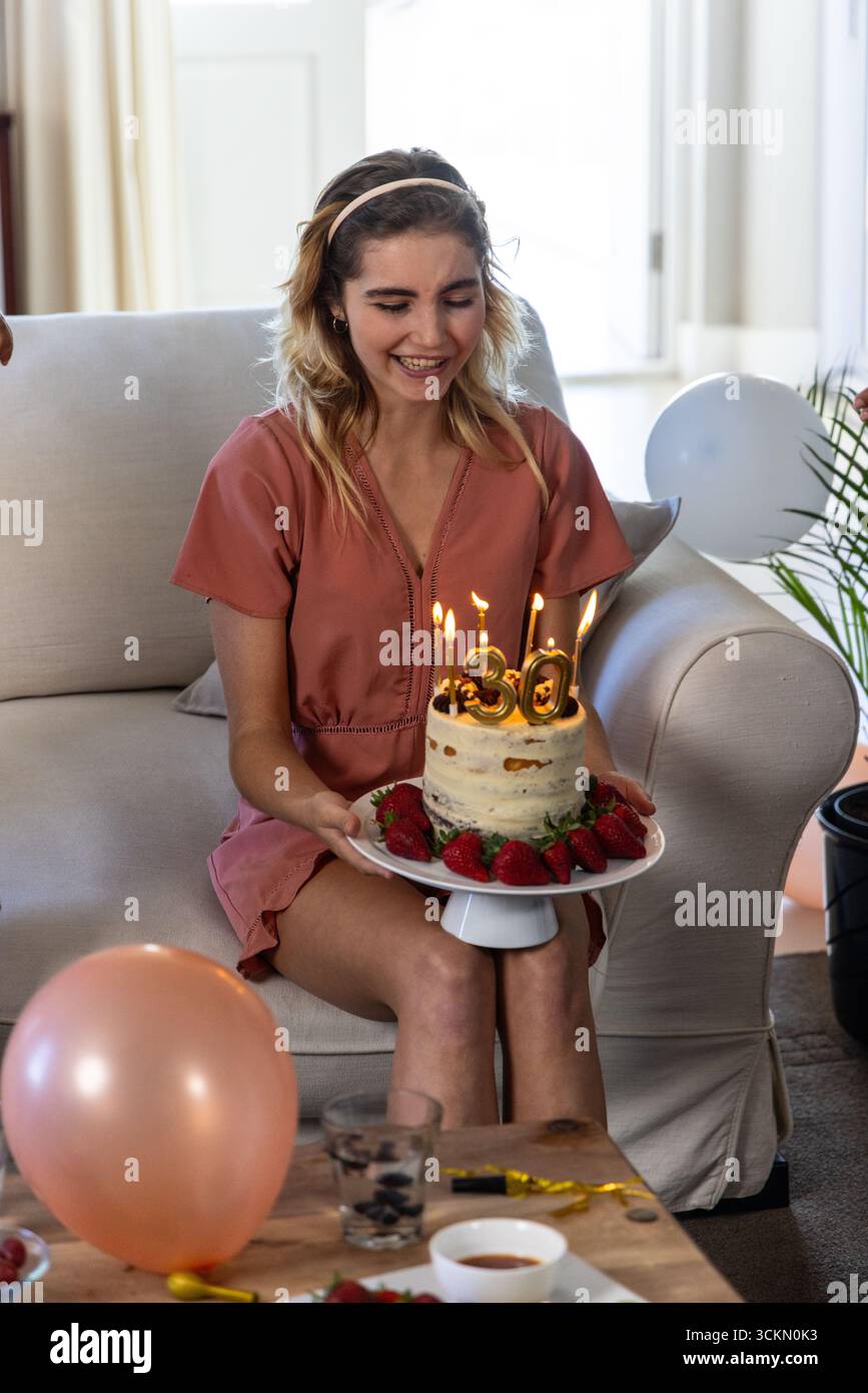 Frau, die zu Hause auf der Couch sitzt und Geburtstagskuchen hält, mit brennenden Kerzen der Nummer 30 auf dem Teller Stockfoto