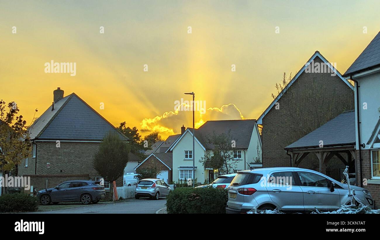 Elsenham, England – 21. Oktober 2024: Ein strahlender Herbstuntergang wirft um 17:47 Uhr Ortszeit goldene Strahlen über den Franklin Drive in Elsenham, Essex. Die Sonne taucht hinter einer Reihe von Wohnhäusern ein und beleuchtet den Himmel mit warmen Tönen, während Lichtstrahlen durch verstreute Wolken brechen. Geparkte Fahrzeuge, saisonales Laub und Halloween-Dekorationen schaffen eine malerische und ruhige Vorstadtszene. Der Moment, der an einem Montagabend festgehalten wird, zeigt den ruhigen Rhythmus des Dorflebens während der wechselnden Jahreszeiten im ländlichen England. Stockfoto