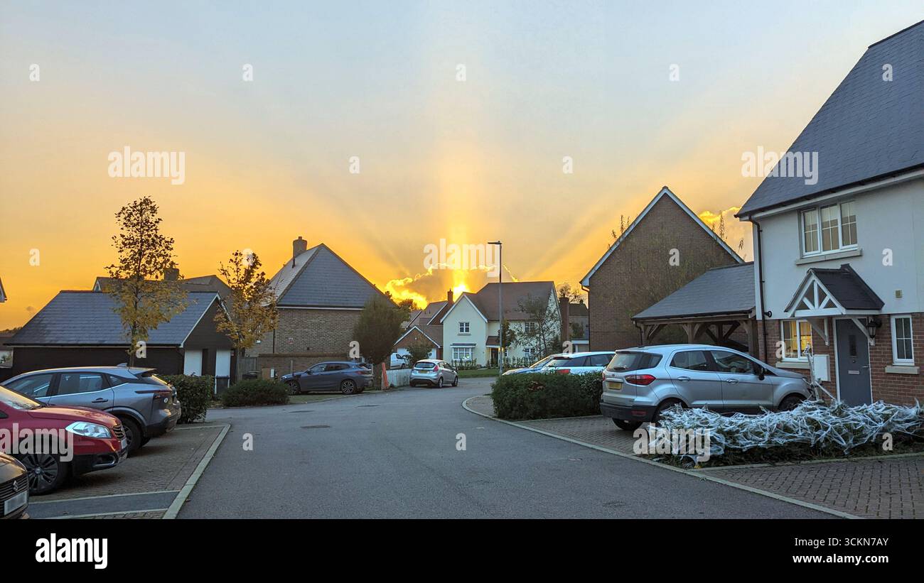 Elsenham, England – 21. Oktober 2024: Ein strahlender Herbstuntergang wirft um 17:47 Uhr Ortszeit goldene Strahlen über den Franklin Drive in Elsenham, Essex. Die Sonne taucht hinter einer Reihe von Wohnhäusern ein und beleuchtet den Himmel mit warmen Tönen, während Lichtstrahlen durch verstreute Wolken brechen. Geparkte Fahrzeuge, saisonales Laub und Halloween-Dekorationen schaffen eine malerische und ruhige Vorstadtszene. Der Moment, der an einem Montagabend festgehalten wird, zeigt den ruhigen Rhythmus des Dorflebens während der wechselnden Jahreszeiten im ländlichen England. Stockfoto