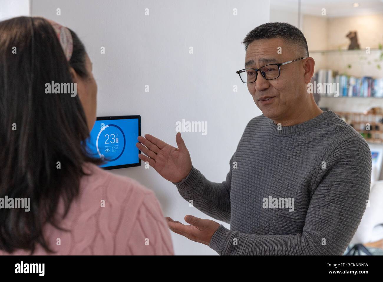 Ein älterer chinesischer Mann mit einer Frau diskutiert intelligente Thermostateinstellungen im Flur zu Hause, Kopierraum Stockfoto