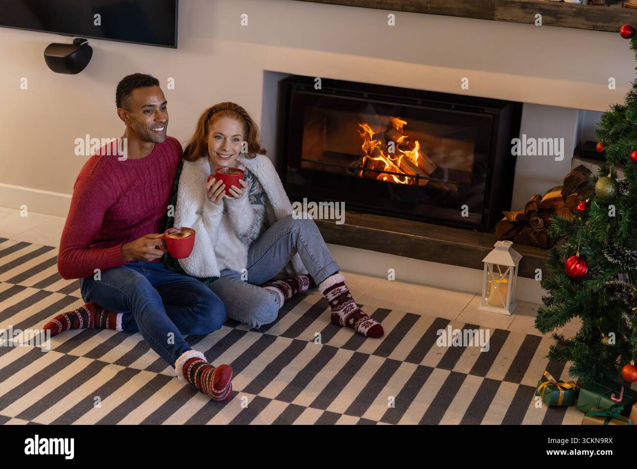 Diverse Paare sitzen auf gestreiften Teppichen vor dem Kamin am weihnachtsbaum mit roten Tassen Stockfoto