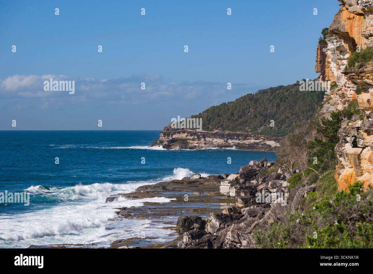 Felsige Klippen, die sich an der Küste mit sanften Meereswellen und grüner Vegetation umarmen, an einem hellen, wolkenbedeckten Tag im Sonnenlicht getaucht. Eine ideale Szene r Stockfoto