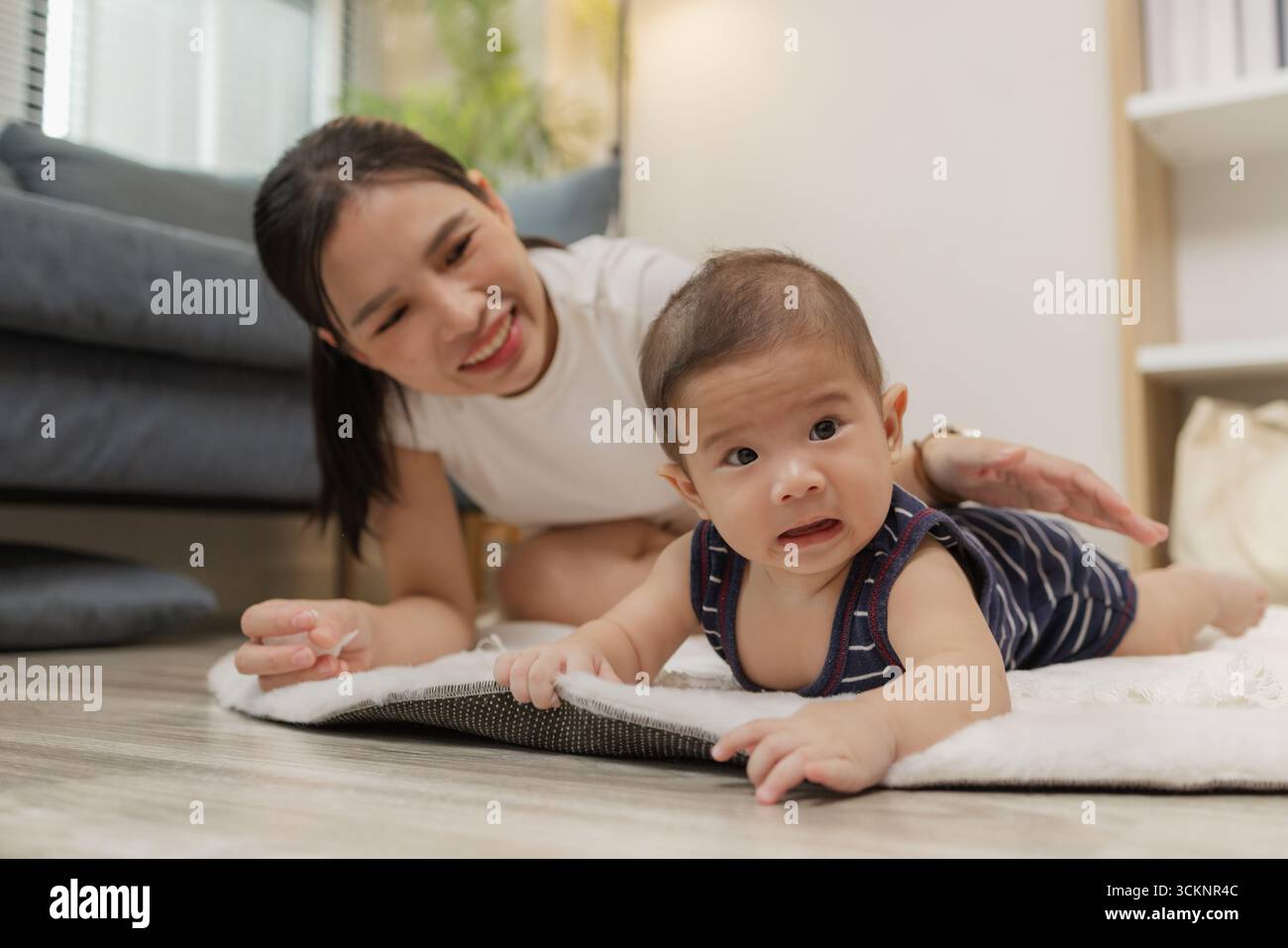 Ein Baby genießt die Bauchzeit auf einer weichen Matte, während die Mutter nah bleibt und es mit einem warmen Lächeln ermutigt. Dieser zärtliche Moment hebt frühe Entwicklungskräfte hervor Stockfoto