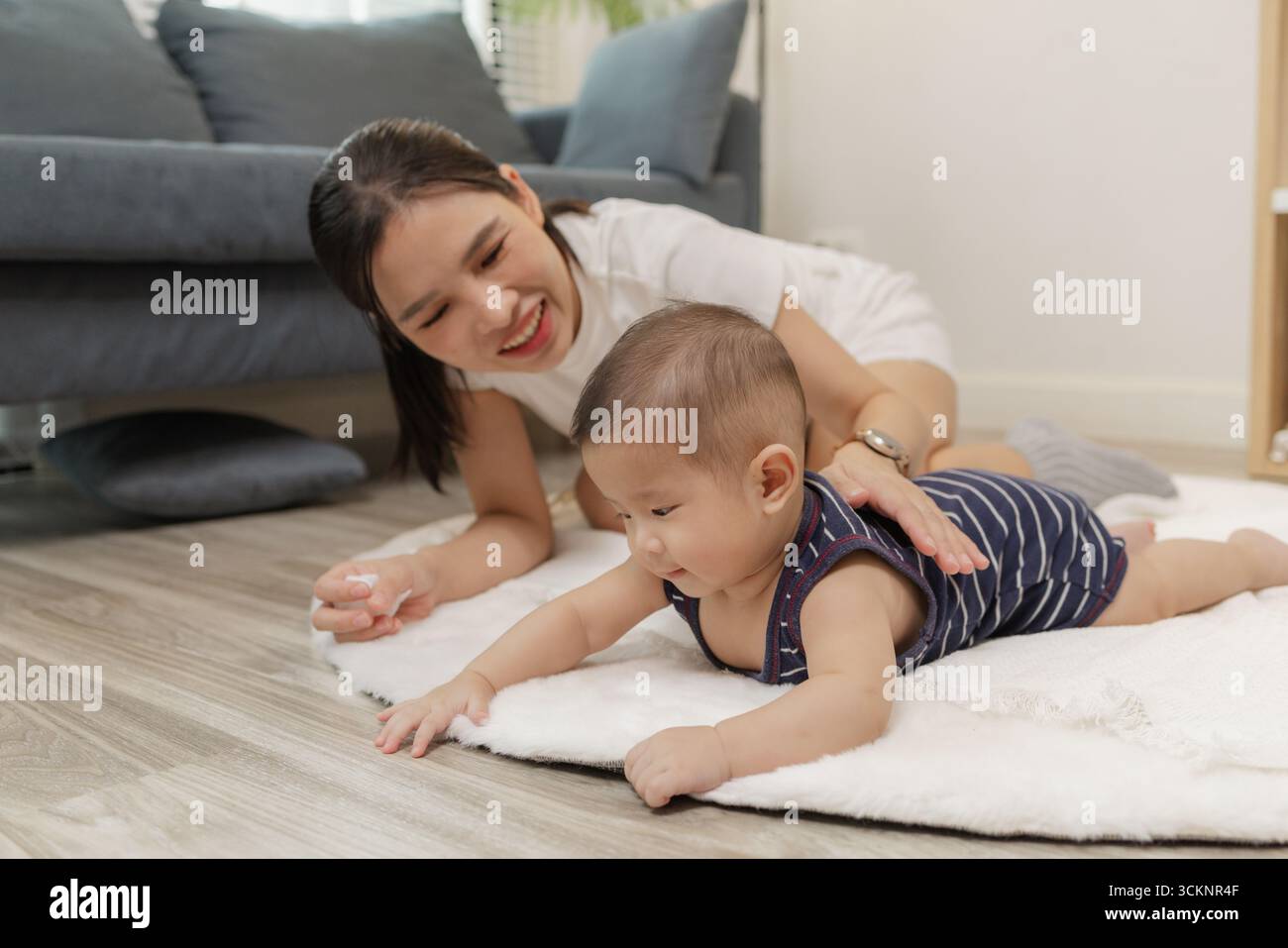 Ein Baby genießt die Bauchzeit auf einer weichen Matte, während die Mutter nah bleibt und es mit einem warmen Lächeln ermutigt. Dieser zärtliche Moment hebt frühe Entwicklungskräfte hervor Stockfoto