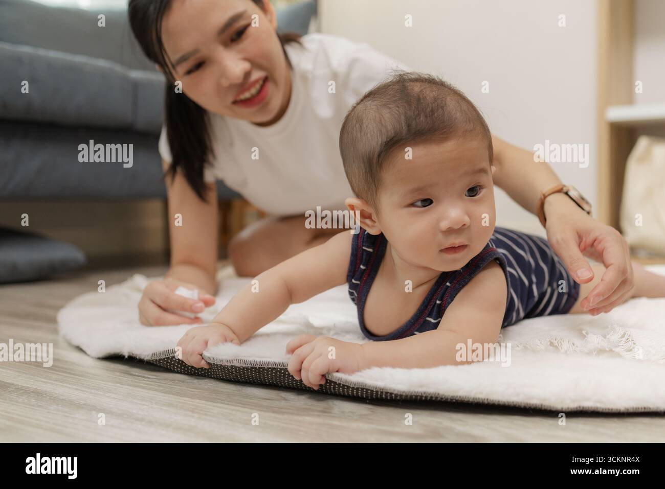 Ein Baby genießt die Bauchzeit auf einer weichen Matte, während die Mutter nah bleibt und es mit einem warmen Lächeln ermutigt. Dieser zärtliche Moment hebt frühe Entwicklungskräfte hervor Stockfoto